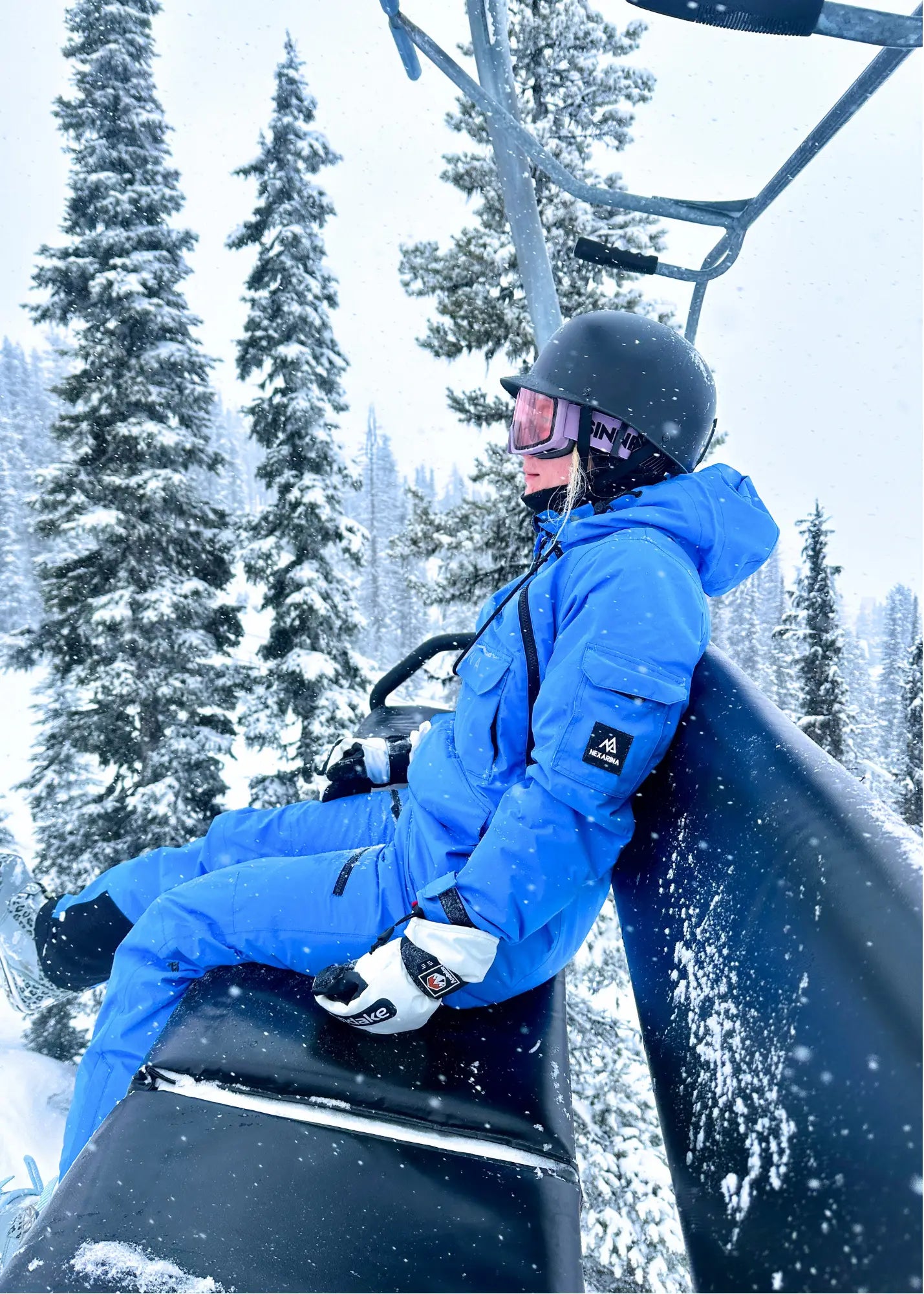 Person in a blue snowsuit sitting on a ski lift with snowy trees in the background