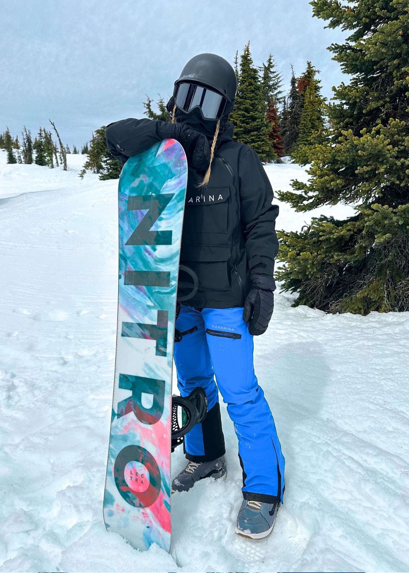 Person holding a Nitro snowboard in a snowy landscape with trees.