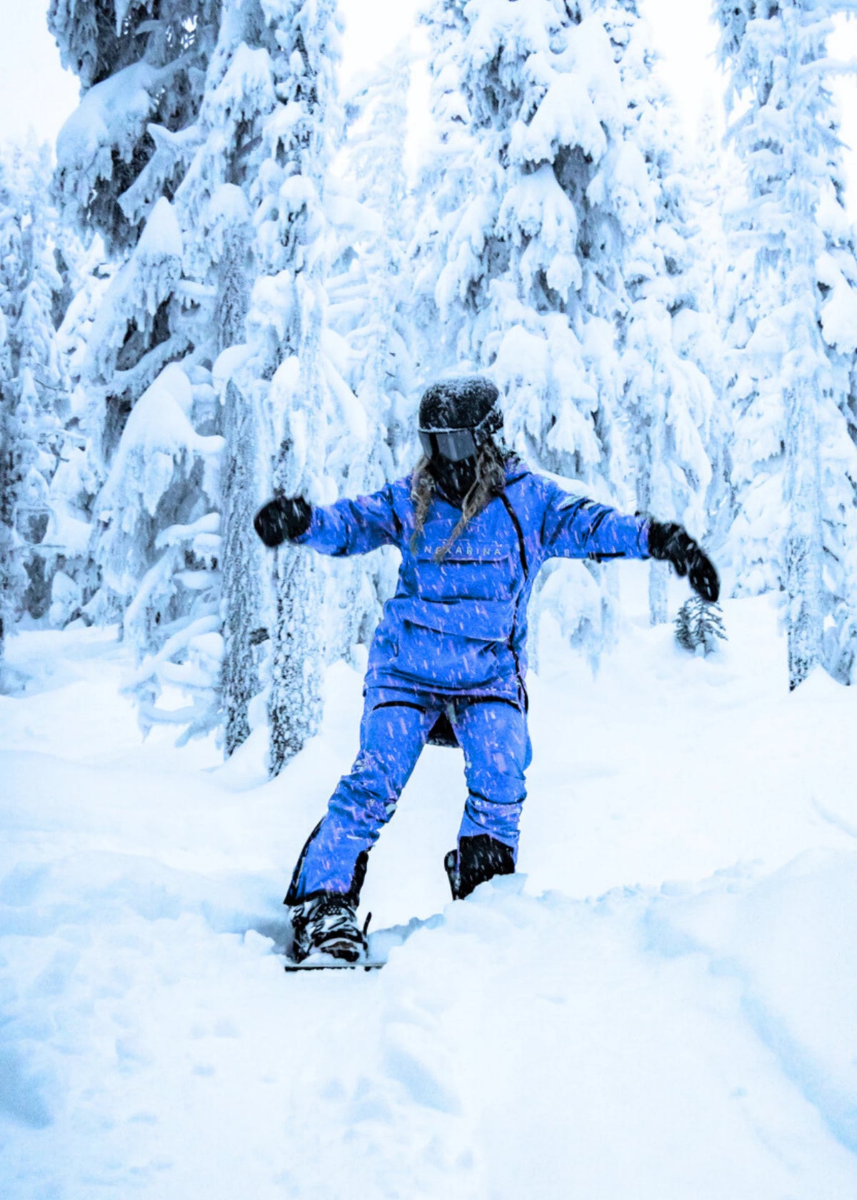 Snowboarder wearing Nexarina Avani Arctic Blue insulated snow pants riding through deep powder snow among snow-covered trees.