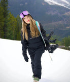 Female snowboarder carrying snowboard while wearing Nexarina black outerwear set in the mountains.