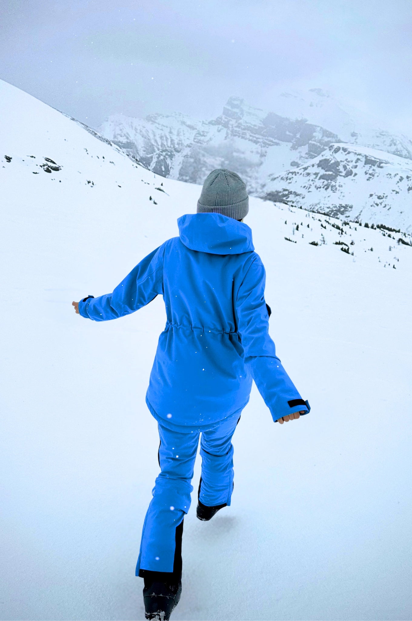 A model walks through the snow in blue Avani snow pants, featuring an adjustable waist, insulation, and flexible design.