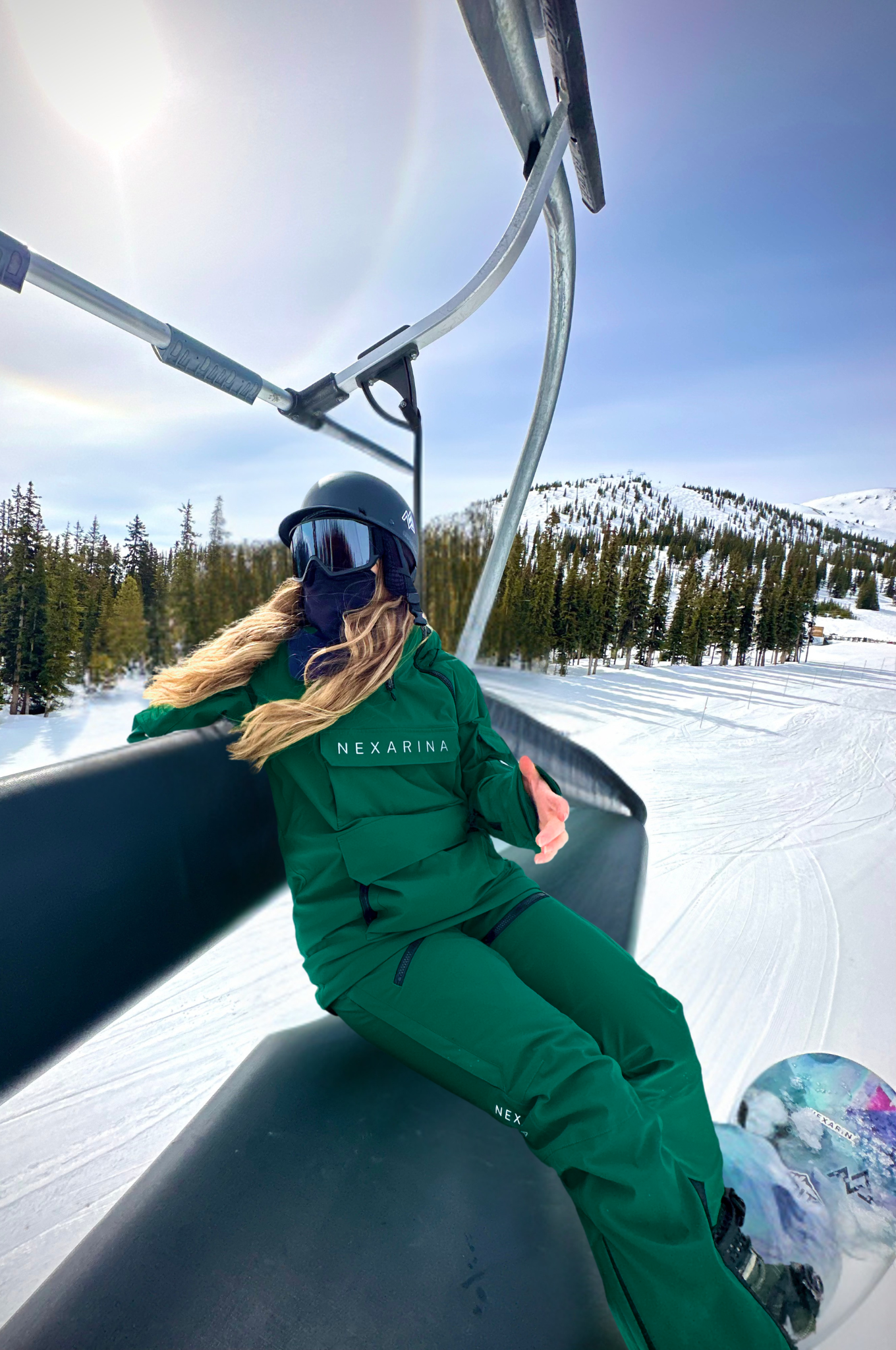 Female snowboarder in Nexarina Avani Hunter Green Snow Pant. She's sitting on a ski lift with snow-covered mountains and trees in the background.