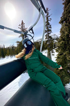 Snowboarder wearing Nexarina Avani green snow pants and Indy jacket, sitting on a ski lift with a snowy mountain landscape in the background. 