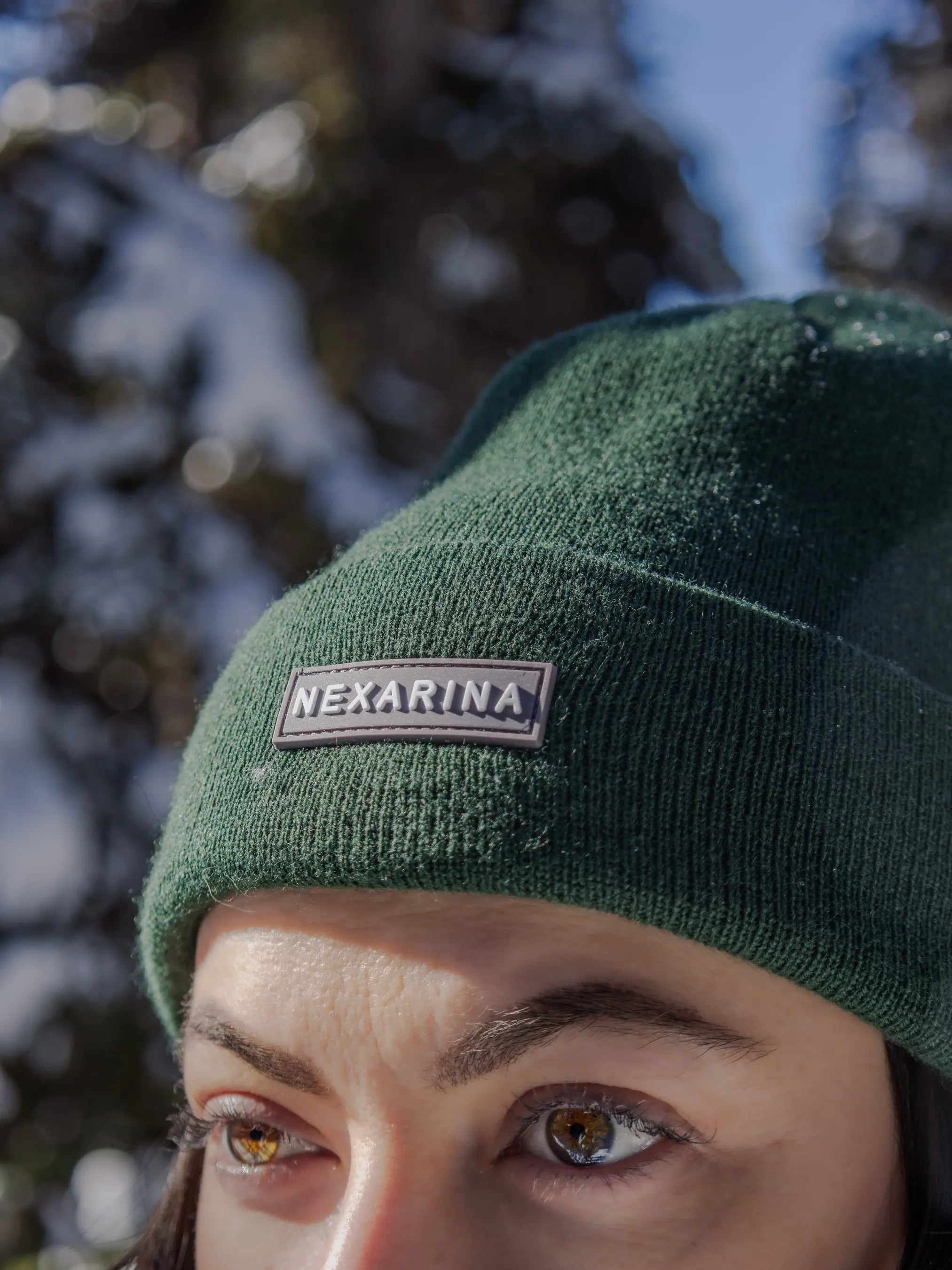 Close-up of a forest green Nexarina beanie with logo patch, worn outdoors with snow-covered trees in the background.