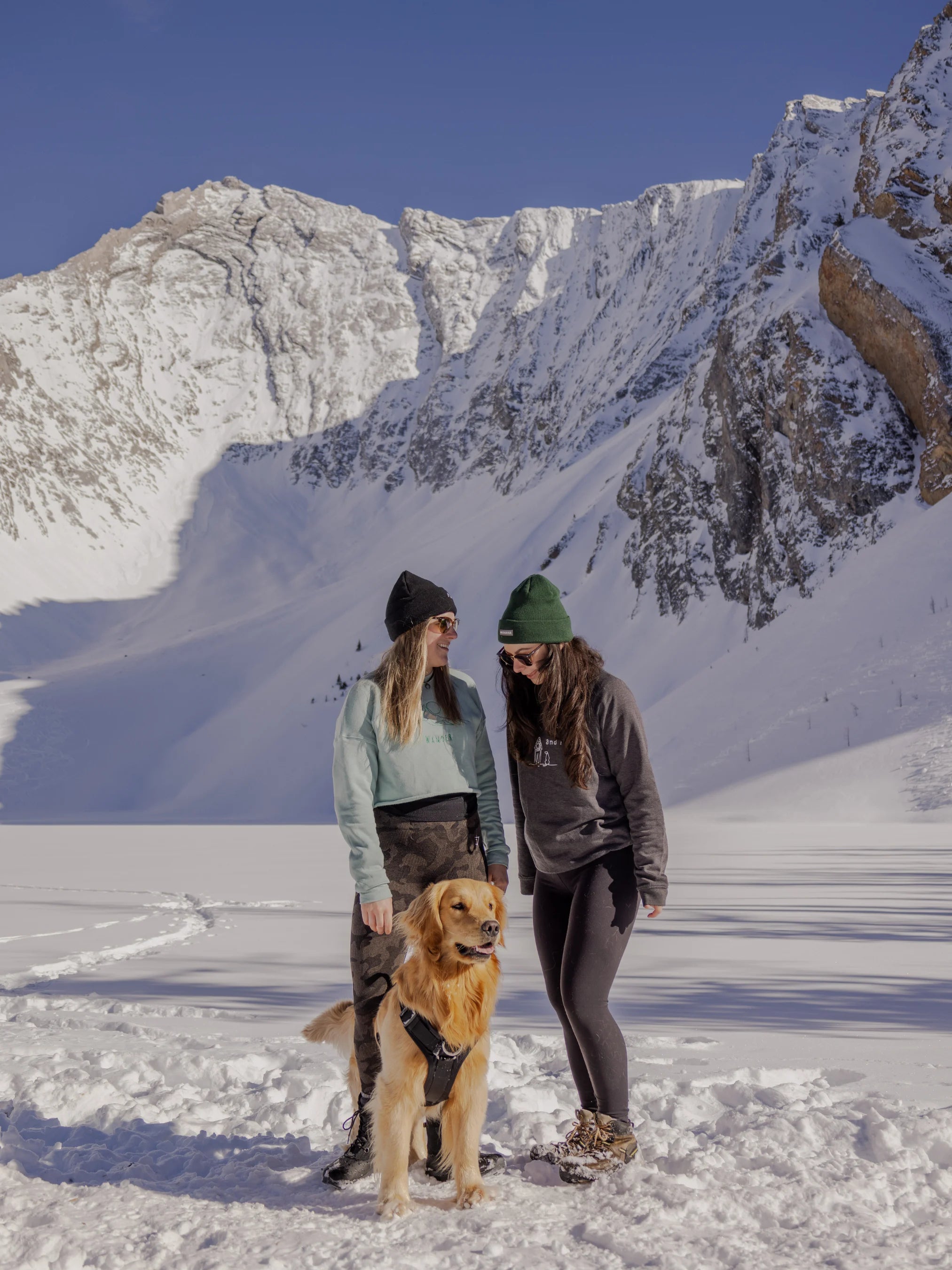 Two women wearing Nexarina gear and beanies, standing in the snow with a golden retriever in front of a dramatic alpine backdrop.