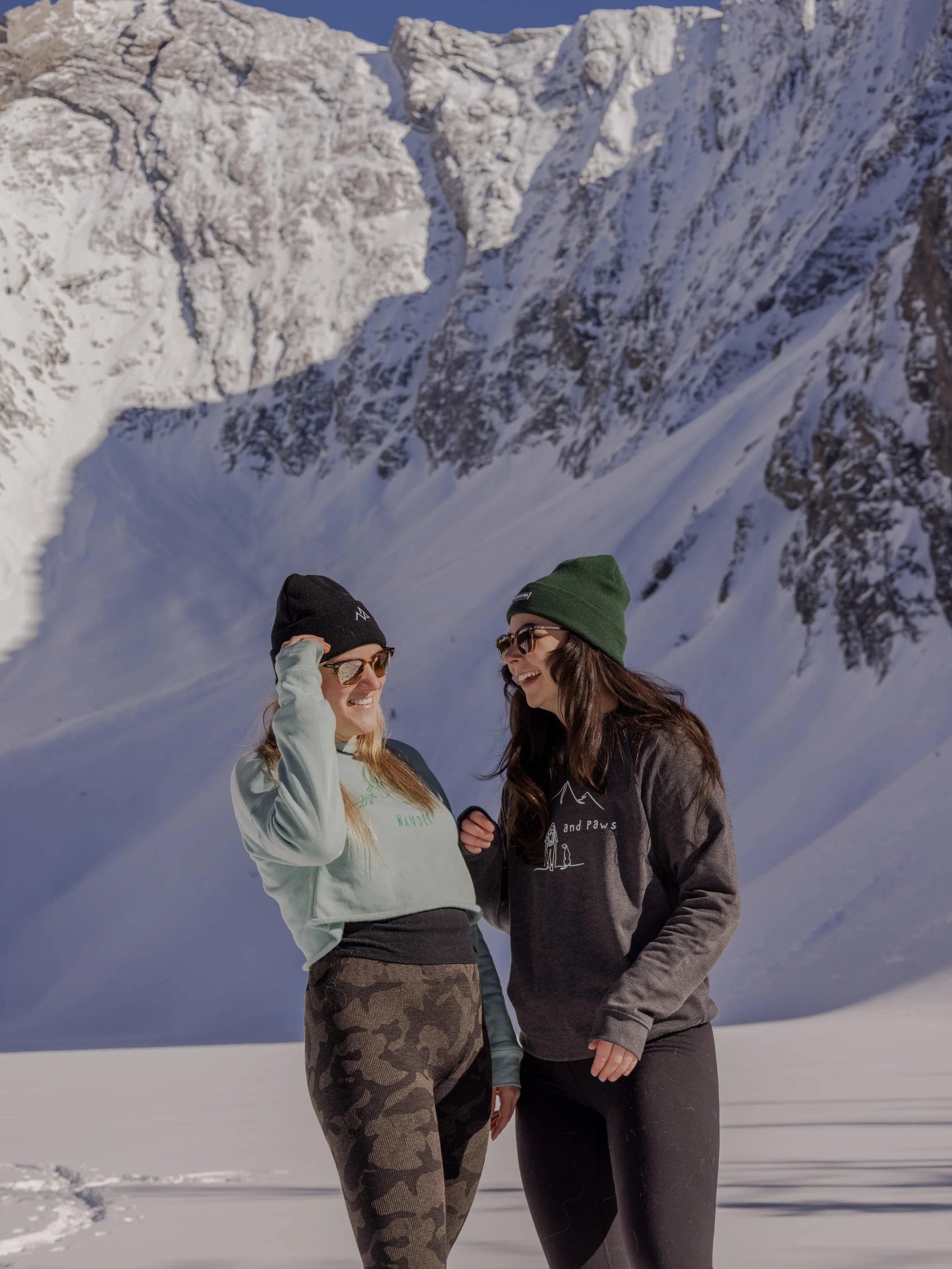 Two women in snow boots and Nexarina gear standing in a snowy valley, laughing with the mountains behind them.