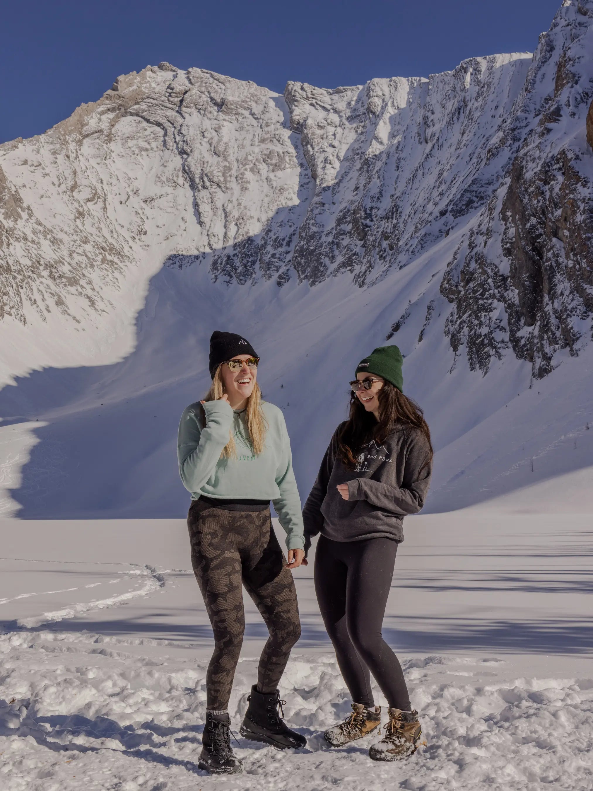 Two women in Nexarina winter apparel and beanies laughing in front of a steep snowy mountain face.