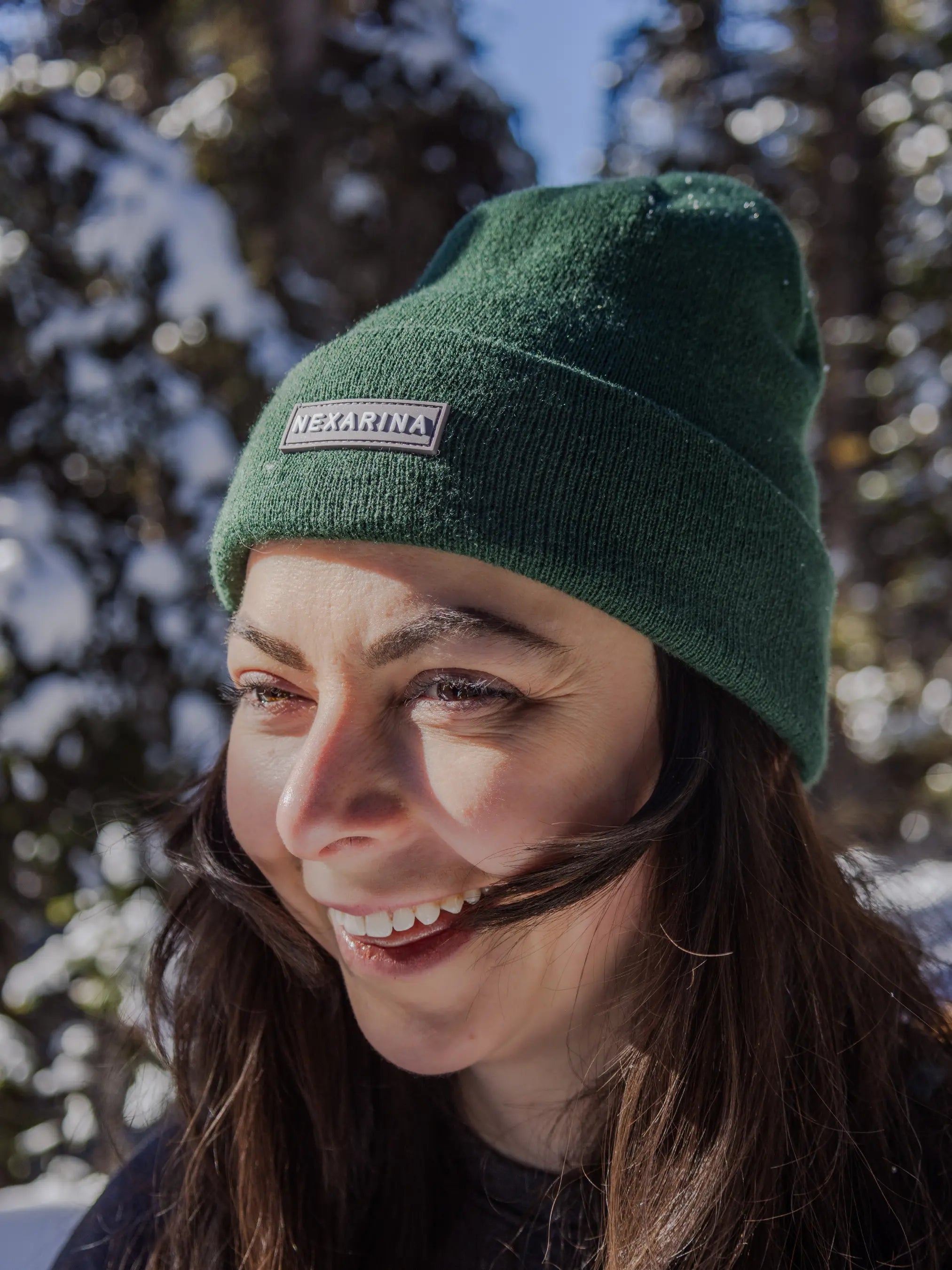 Close-up of a smiling woman in a Nexarina beanie, with trees and sunshine in the background. Her hair is catching the light.