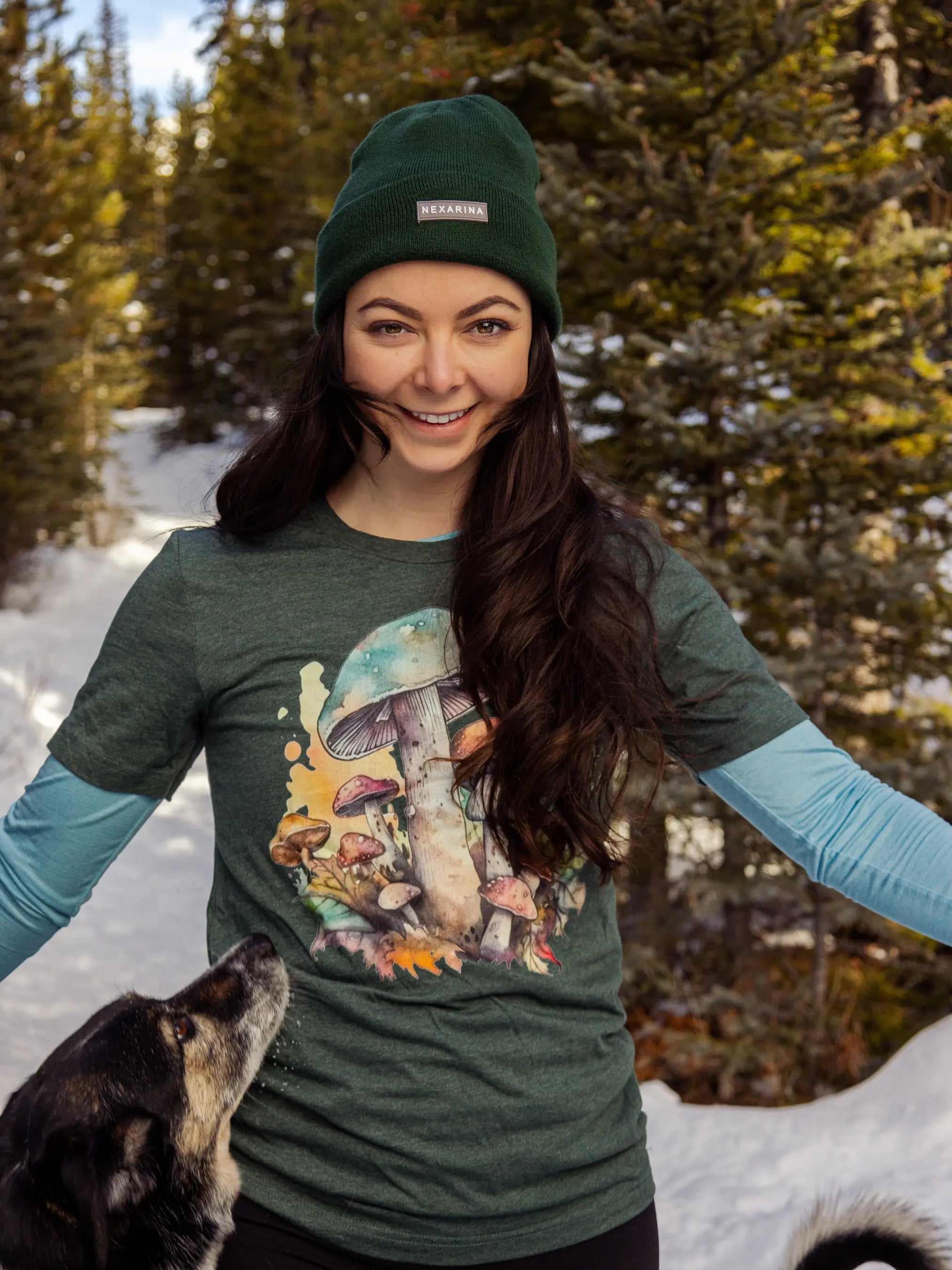 Woman wearing a forest green Nexarina beanie and a mushroom graphic tee, smiling on a snowy trail with a dog looking up at her.