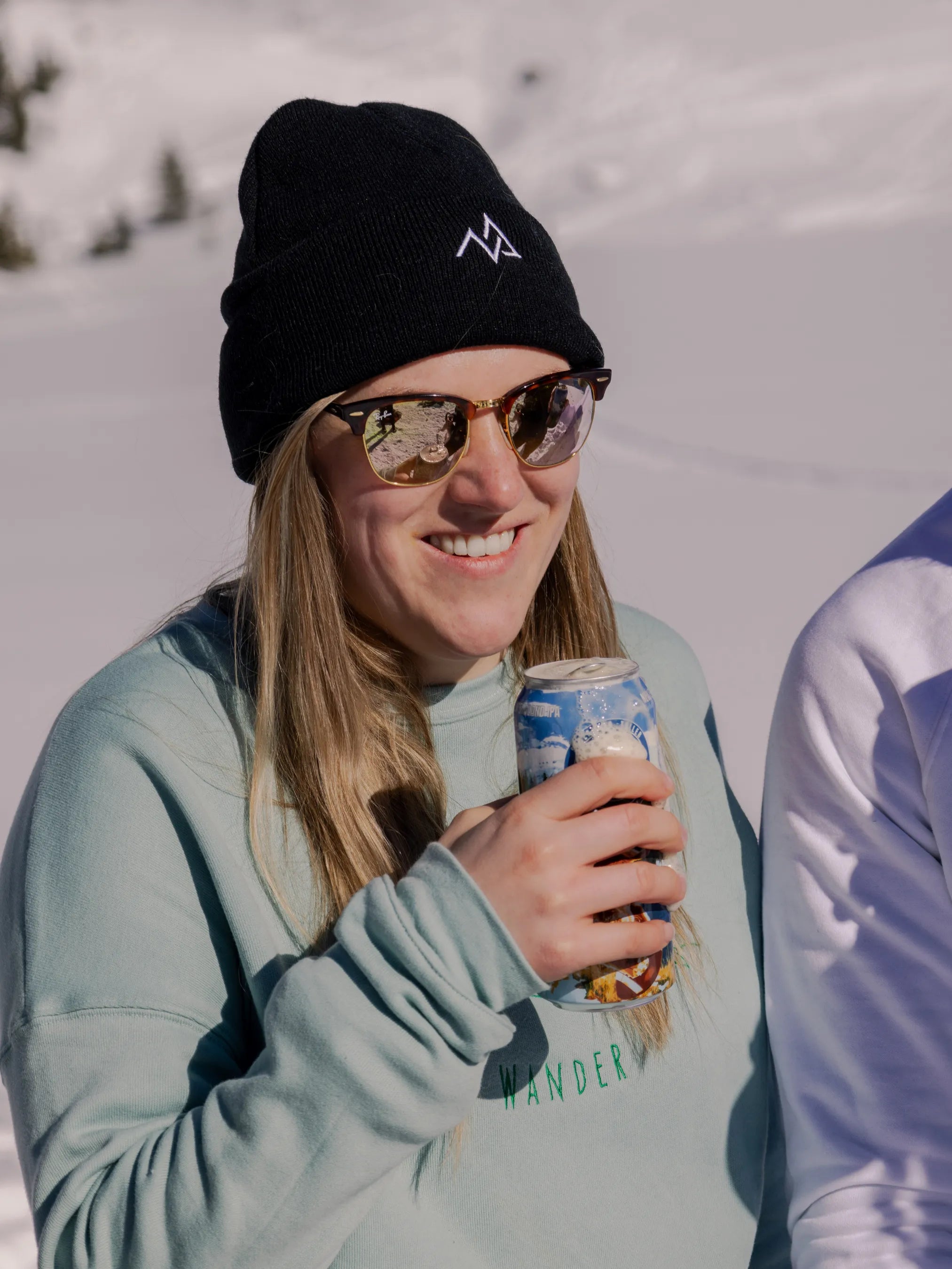 Woman wearing a black Nexarina beanie with embroidered logo, smiling in the sun with snowy mountains behind her.