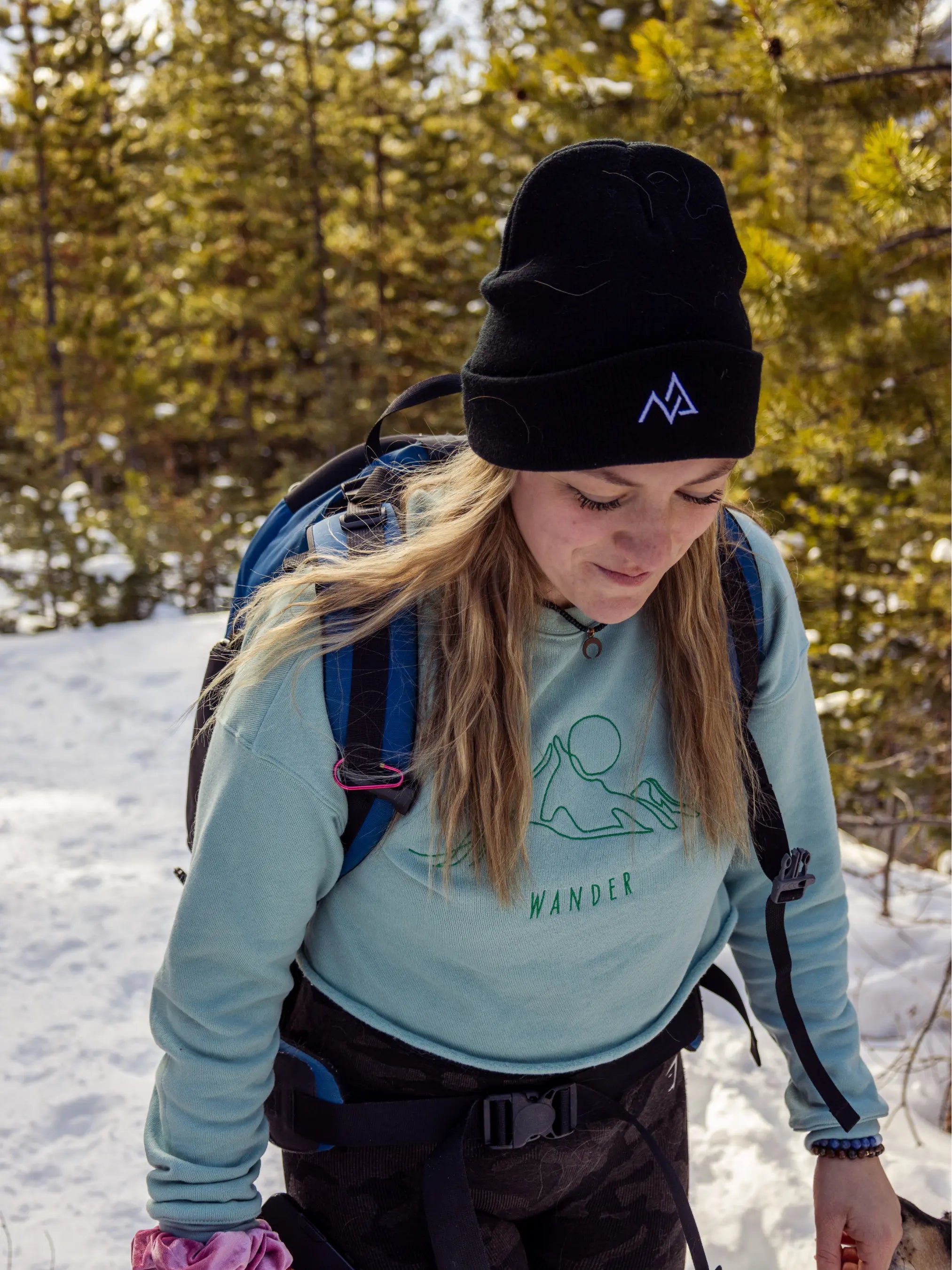 Close-up of a woman hiking in a black Nexarina beanie and mint green crewneck, trekking through a snowy forest.