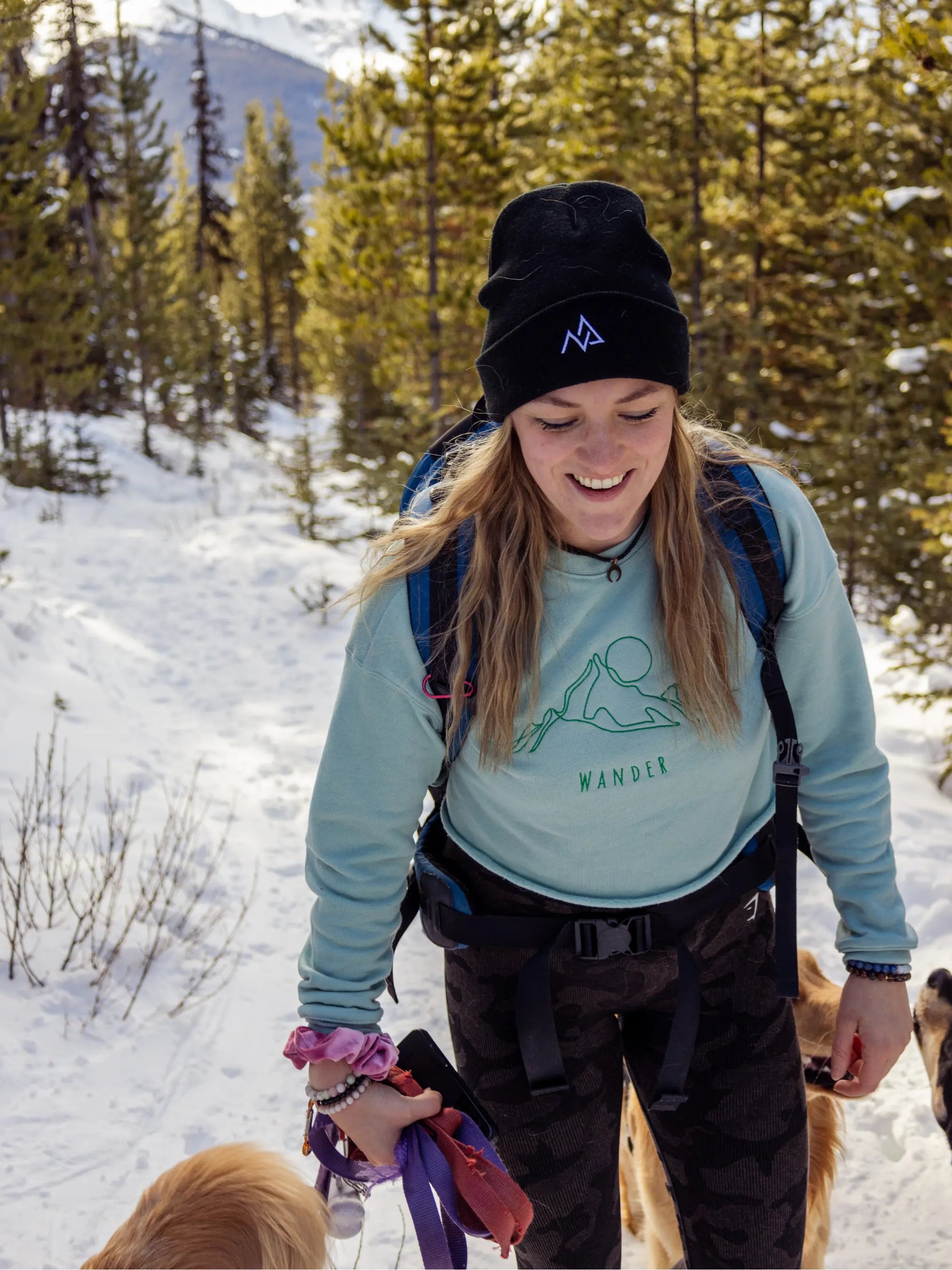 Woman wearing a black Nexarina beanie, smiling while hiking snowy trails with dogs around her.