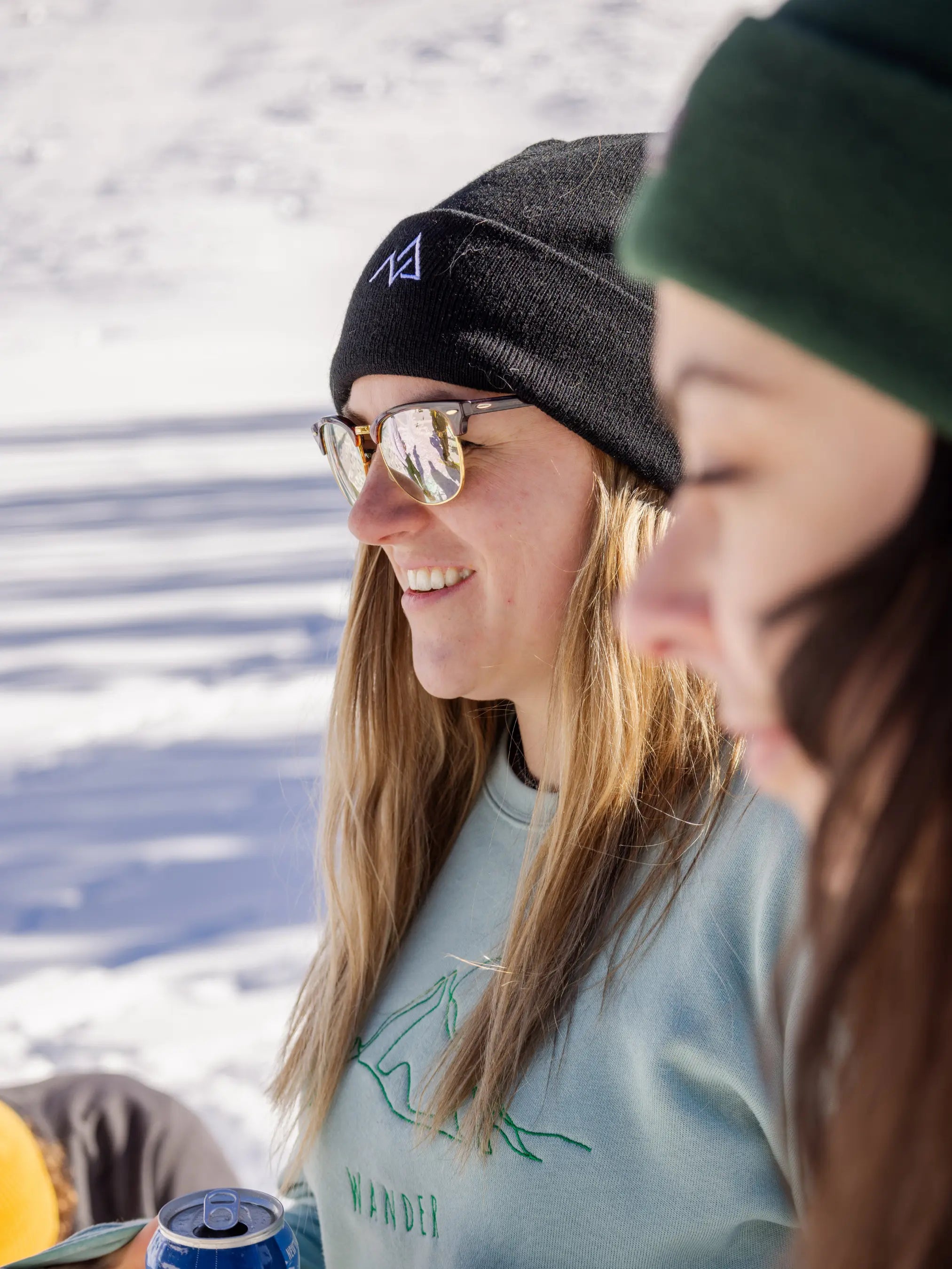 Side profile of a woman in a black Nexarina beanie and reflective sunglasses, holding a drink and smiling outdoors.