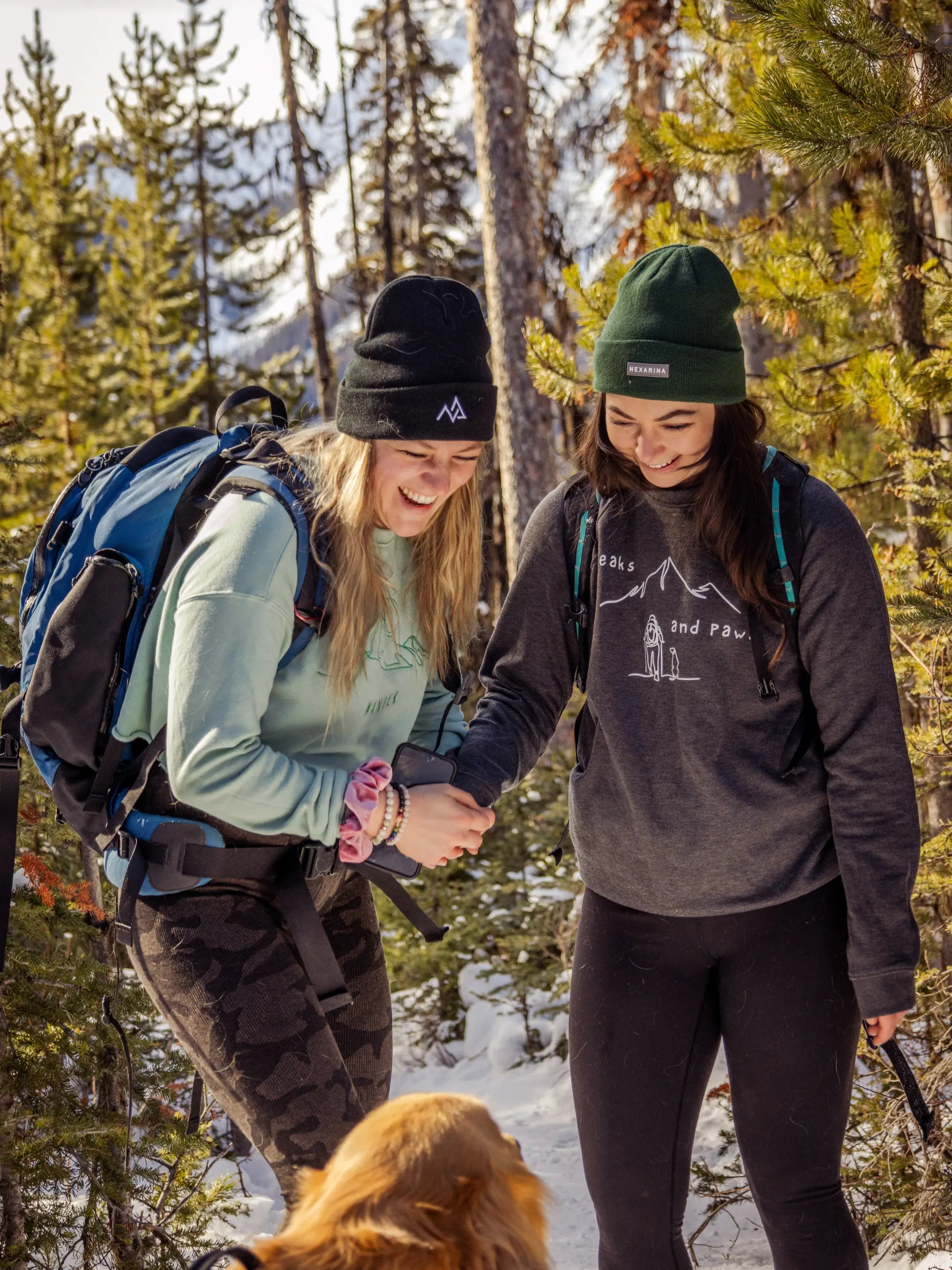 Two women laughing in the forest, one wearing a black Nexarina beanie and the other in green, holding hands and surrounded by snow and pine trees.