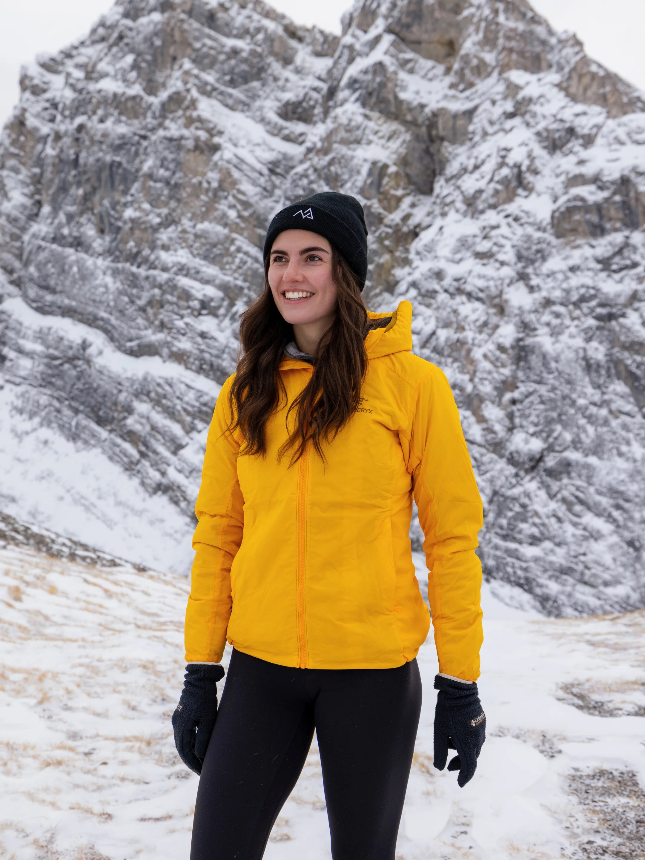 Woman wearing a black Nexarina beanie and bright yellow jacket standing in front of rugged, snow-covered mountain cliffs with a smile.