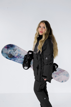 "Side profile of a woman in a black Nexarina snowboard jacket, holding a snowboard with a colorful design, against a white backdrop.