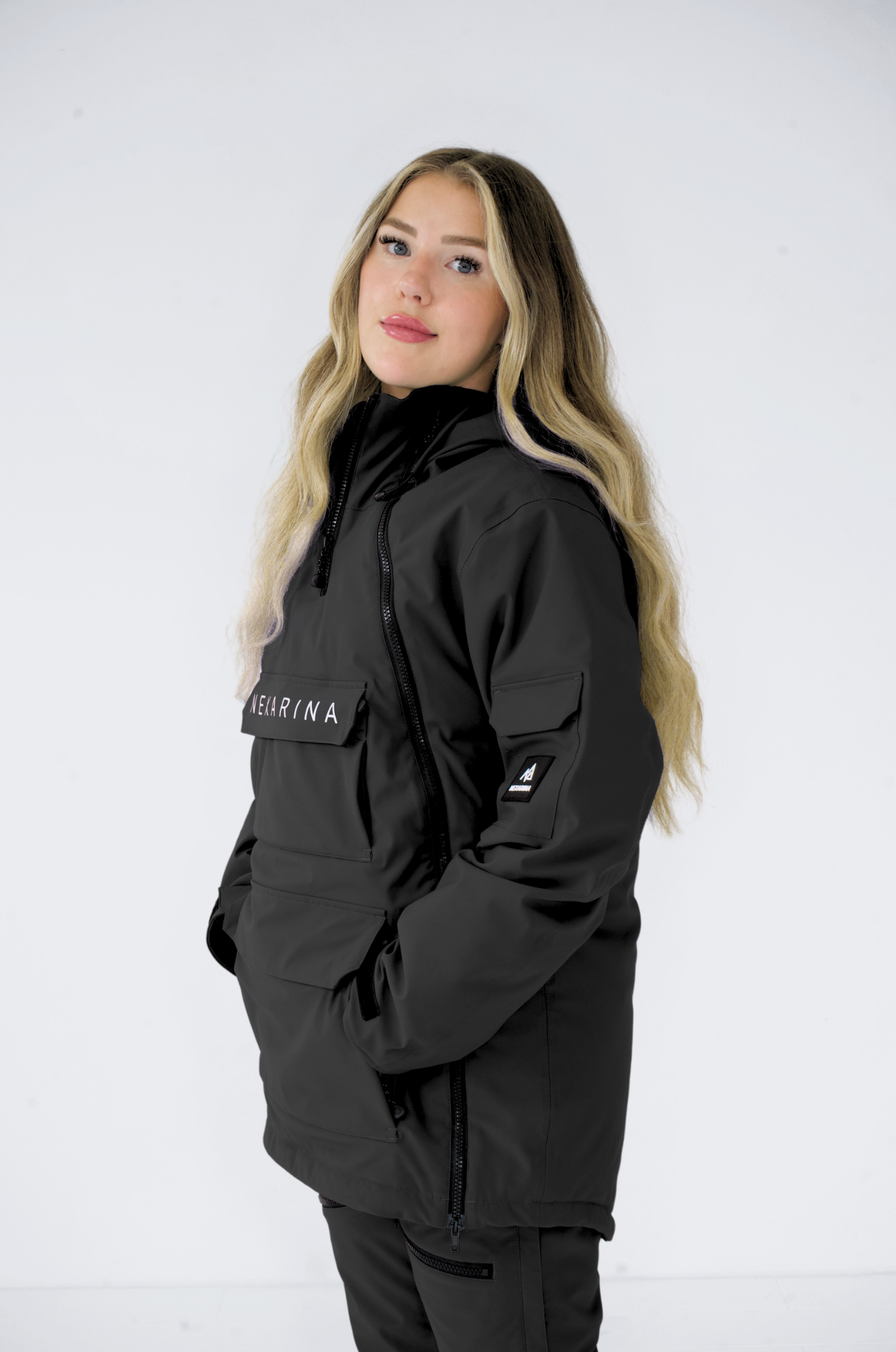 Front view of a woman wearing a black Nexarina snowboard jacket, looking towards the camera, against a white backdrop