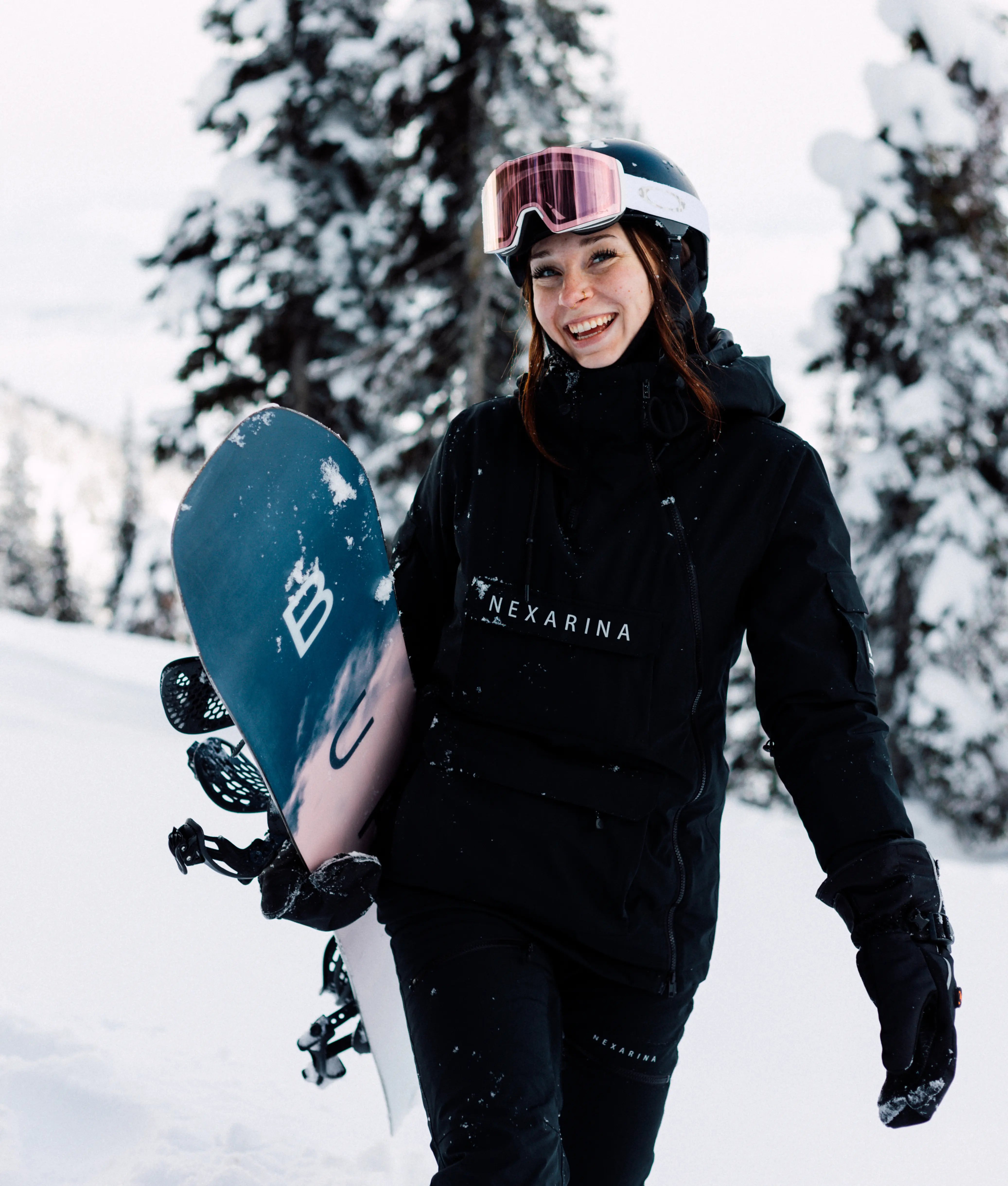Woman smiling while holding snowboard in snowy setting, wearing Nexarina Indy Snow Jacket and pants
