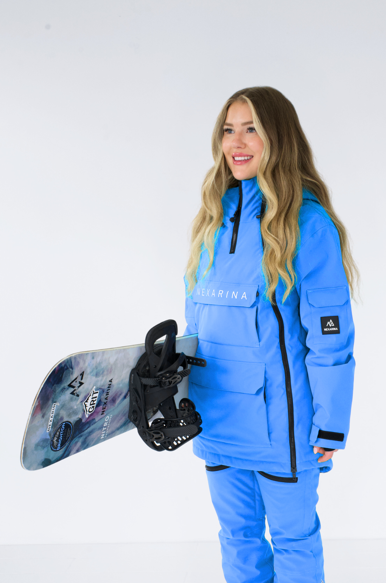 Model in a bright blue Nexarina snowboarding jacket holds a snowboard, smiling warmly, in a studio setting.
