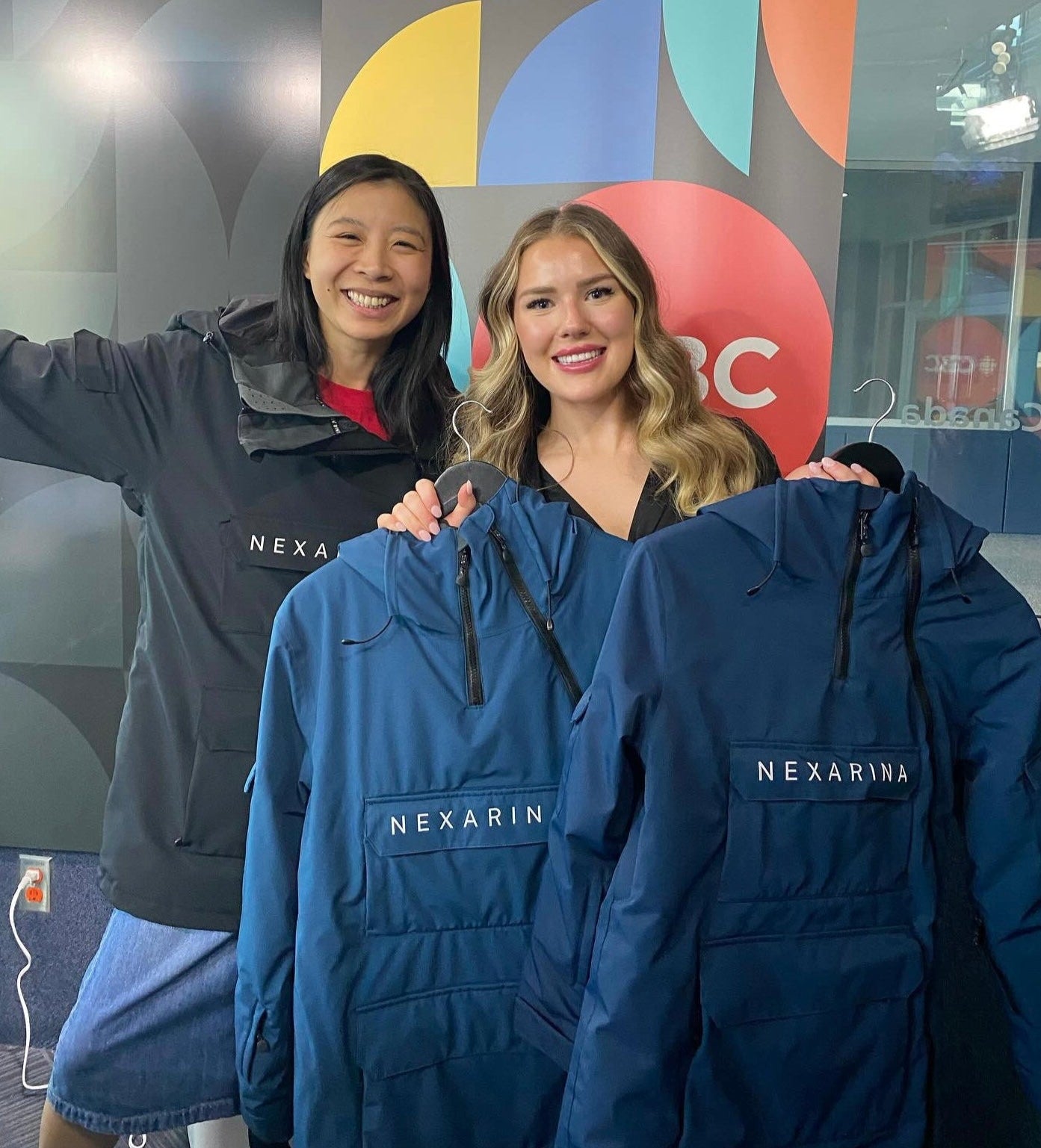 Two women holding blue jackets with 'Nexarin' branding in front of a colorful background.