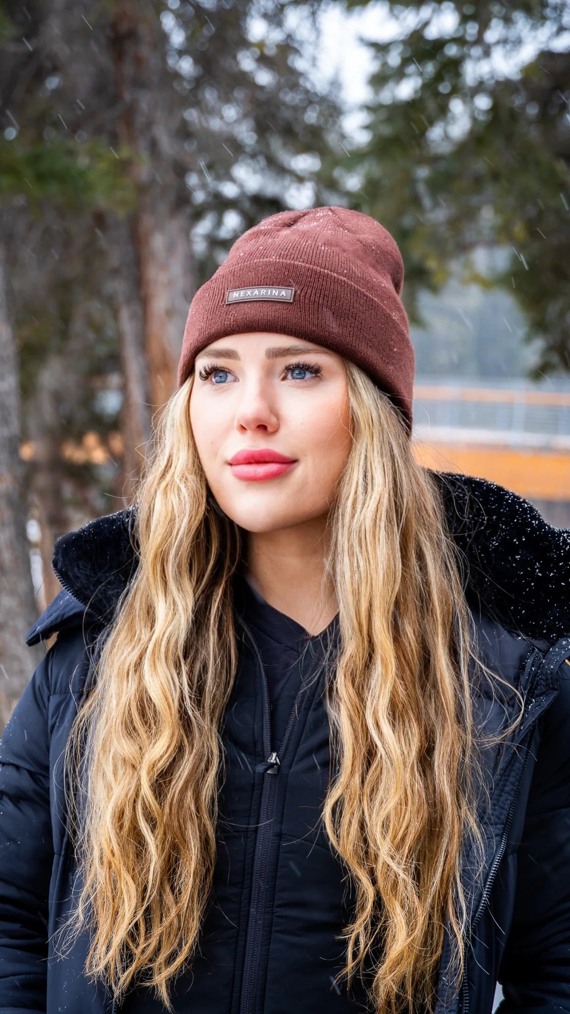 Portrait of a young woman wearing Nexarina Coco beanie, snow falling gently around her.