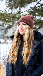 Woman in black jacket and Nexarina Coco brown beanie standing in snowy forest.