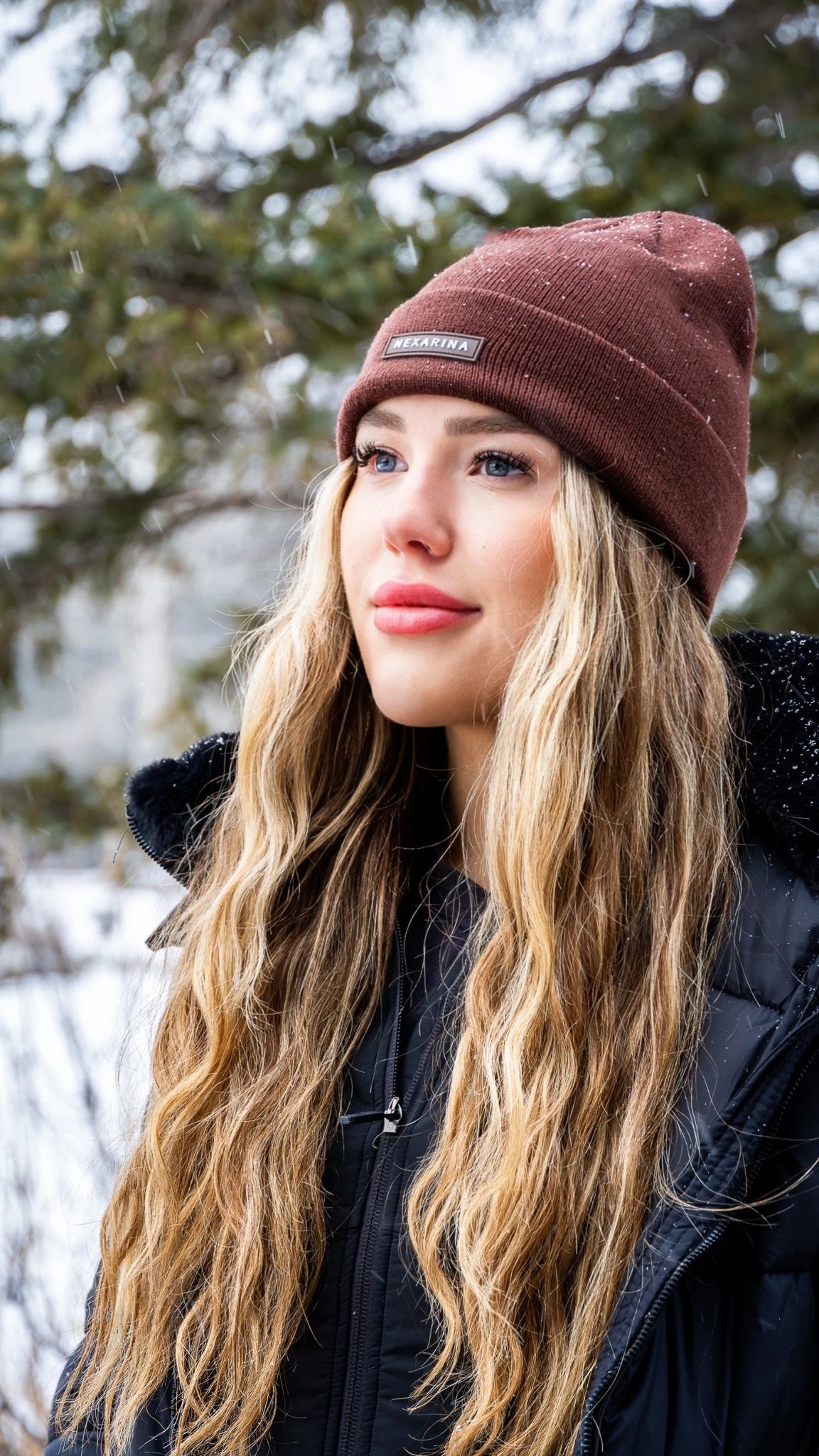 Blonde woman wearing Nexarina Coco beanie in brown, looking thoughtfully in a snowy setting.