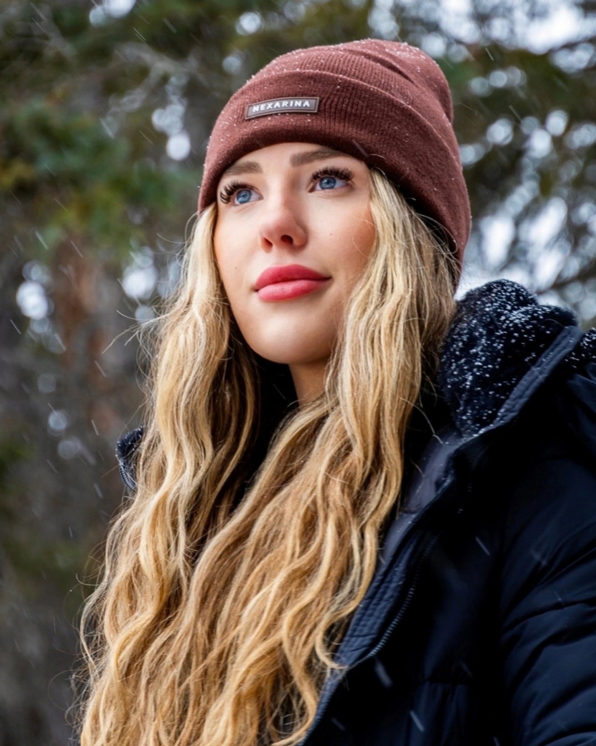 Close-up of a young woman in a Nexarina Coco brown beanie, snow gently falling around her.