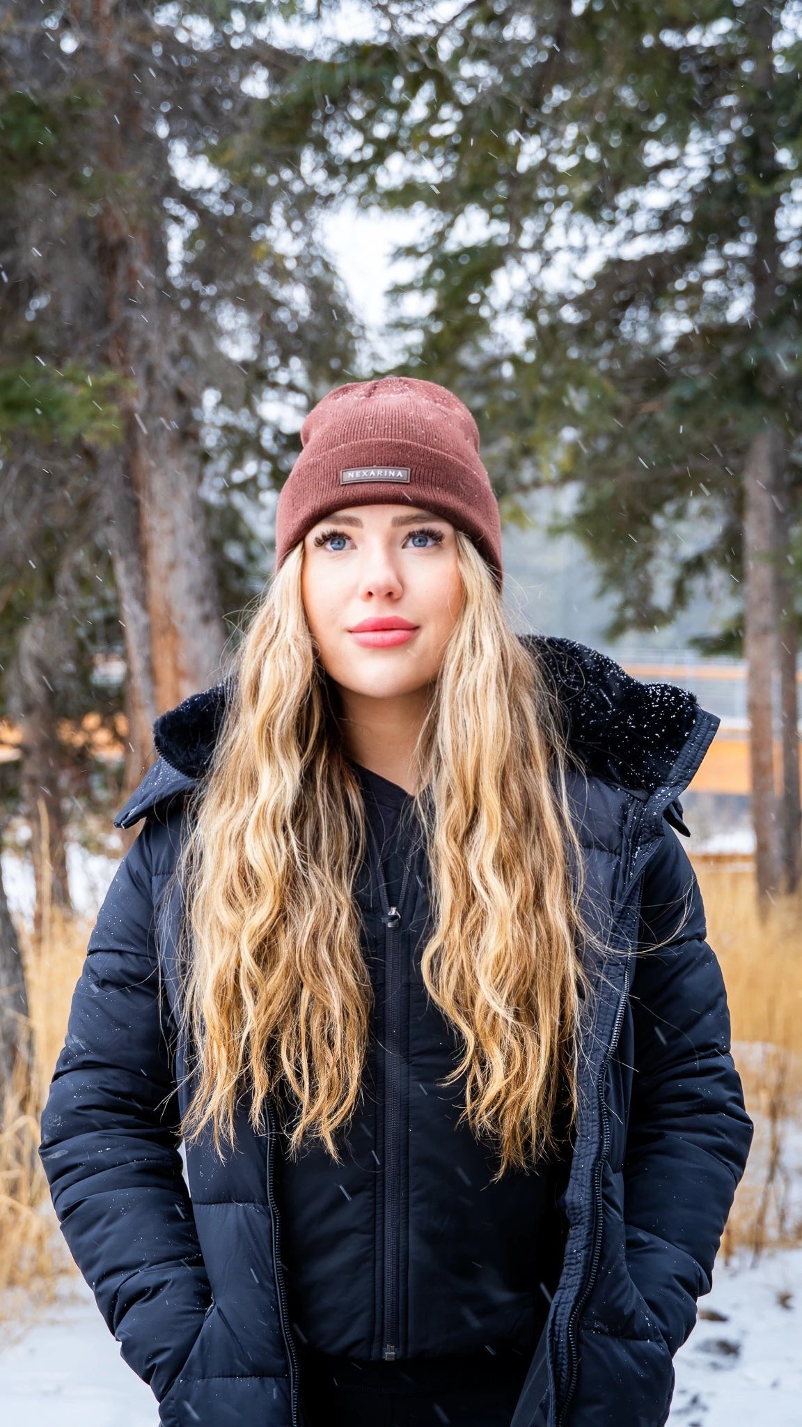 Blonde woman in Nexarina Coco beanie with a thoughtful gaze, snowy trees in the background.