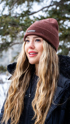 Smiling woman outdoors wearing Nexarina Coco brown beanie, with snow-covered trees in the background.