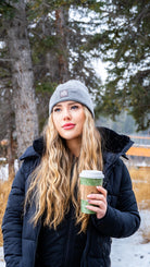 Winter portrait of a woman in a Nexarina grey beanie, enjoying a hot drink in a snowy woodland.