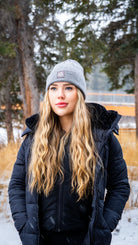 Woman in a grey Nexarina beanie and black coat, smiling gently in a snowy forest setting.