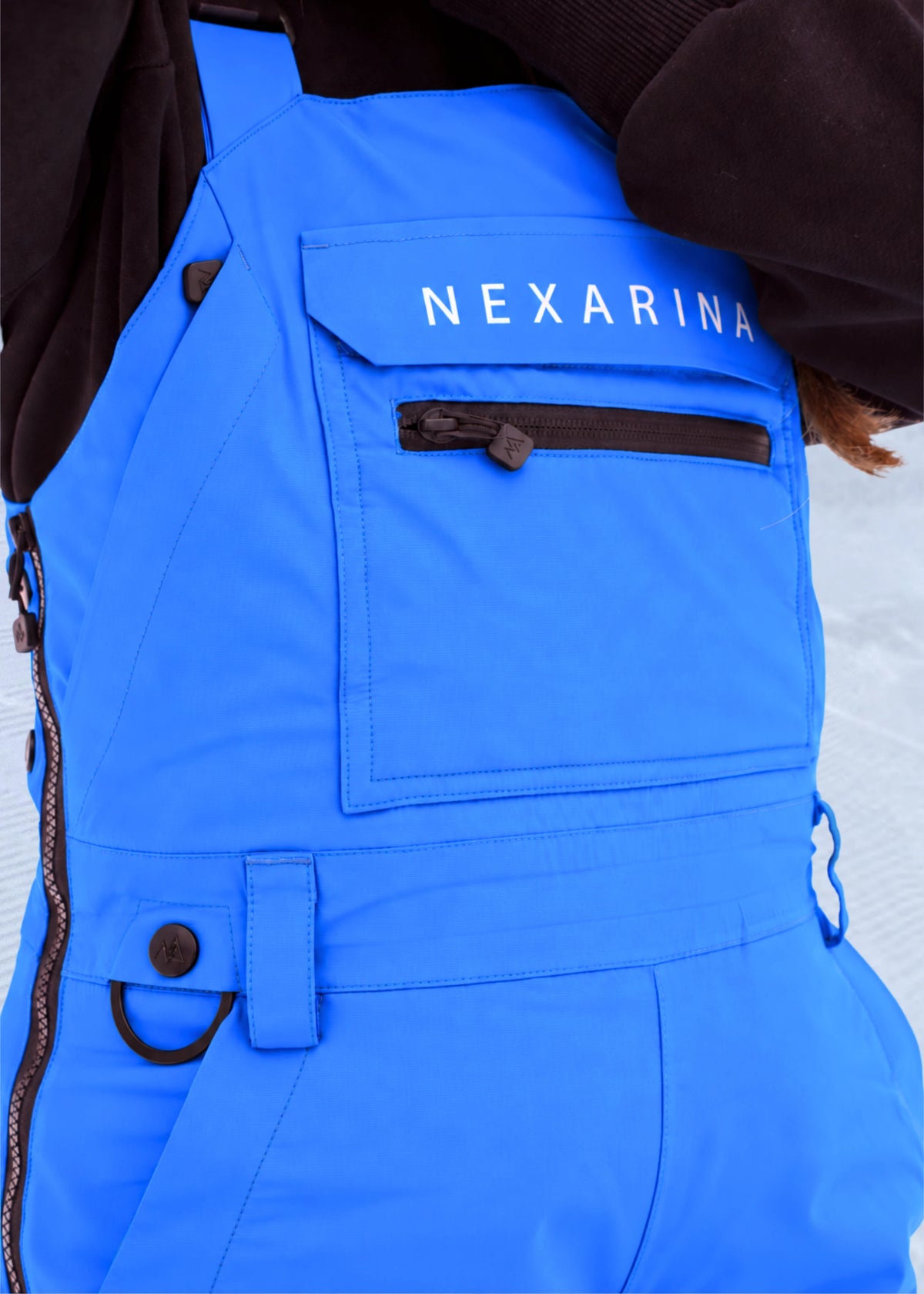 Close-up of Nexarina Everette Arctic Blue snow bibs showing the chest pocket, zipper and Nexarina branding.