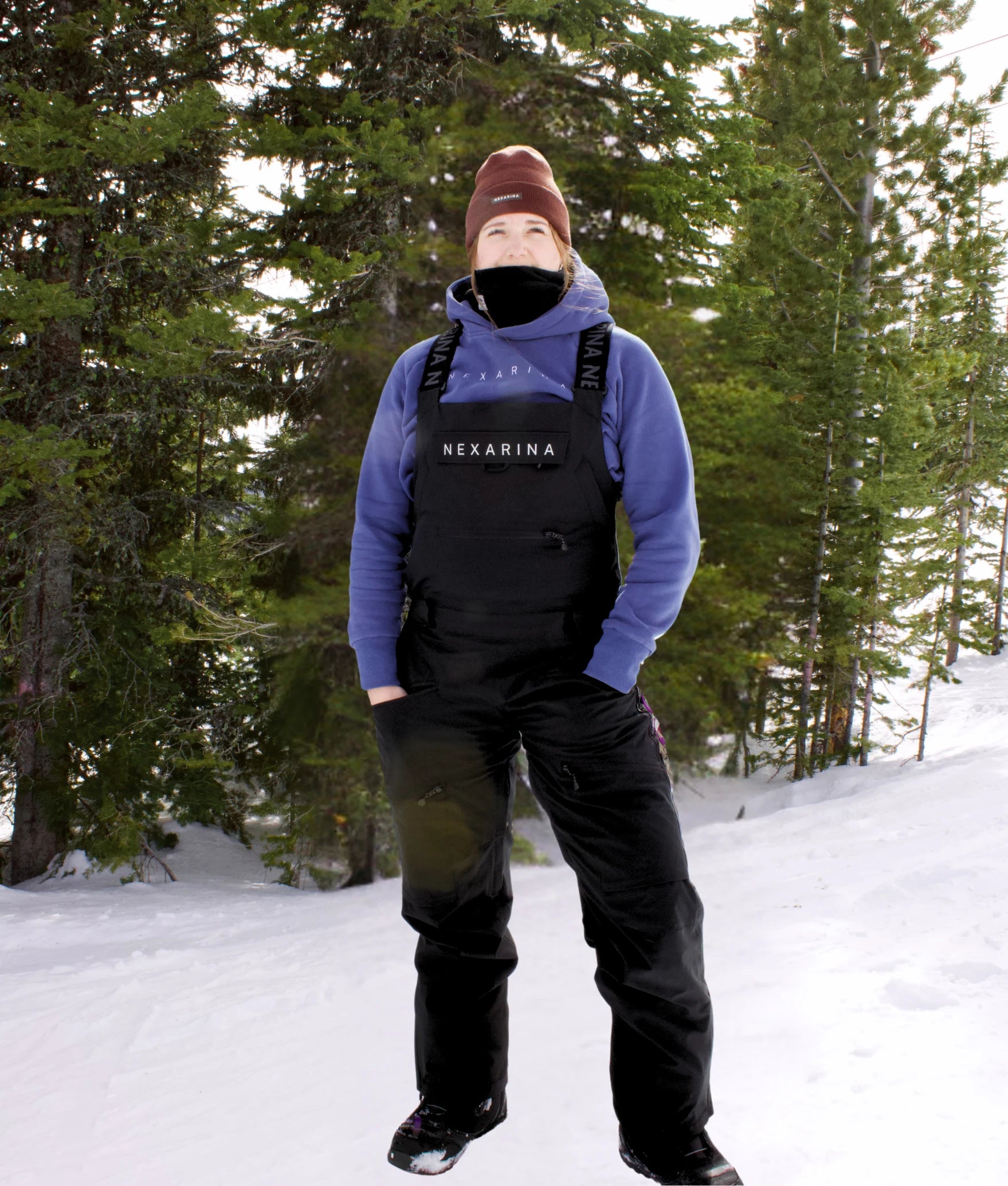 Woman standing in the snow wearing the Nexarina Everette Bib in black, paired with a Nexarina hoodie and the Coco Beanie. The scene shows a forest backdrop with snow-covered ground, highlighting the functional fit and design of Nexarina women’s outerwear.