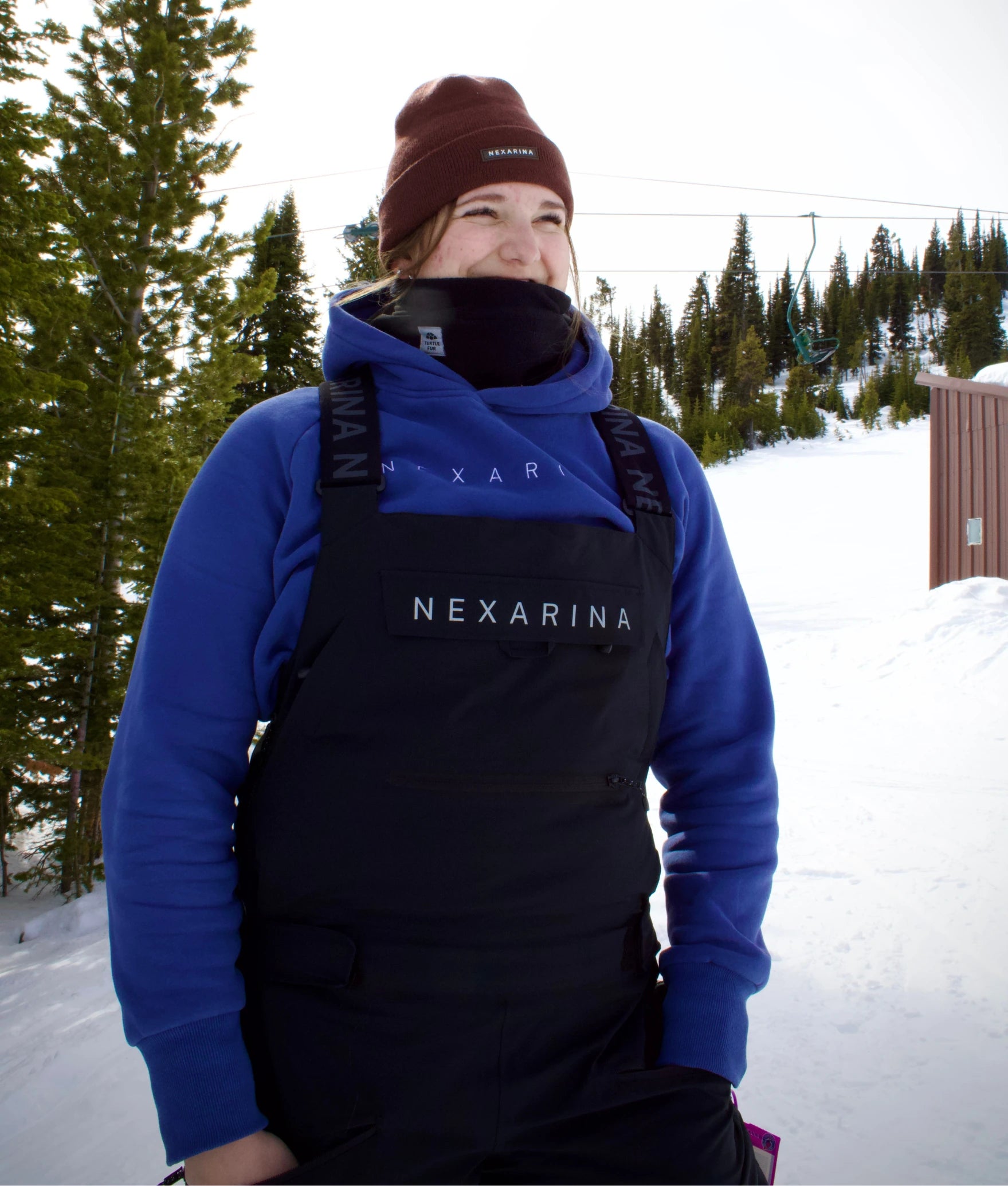 Woman standing in the snow wearing Nexarina Everette Bib black snowboard overall.