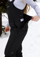 Woman demonstrating the side-opening drop seat zipper on Nexarina Everette Classic Black women's snow bibs.