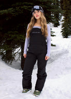 Woman wearing Nexarina Everette Classic Black insulated snow bibs with adjustable shoulder straps in a snowy mountain setting.