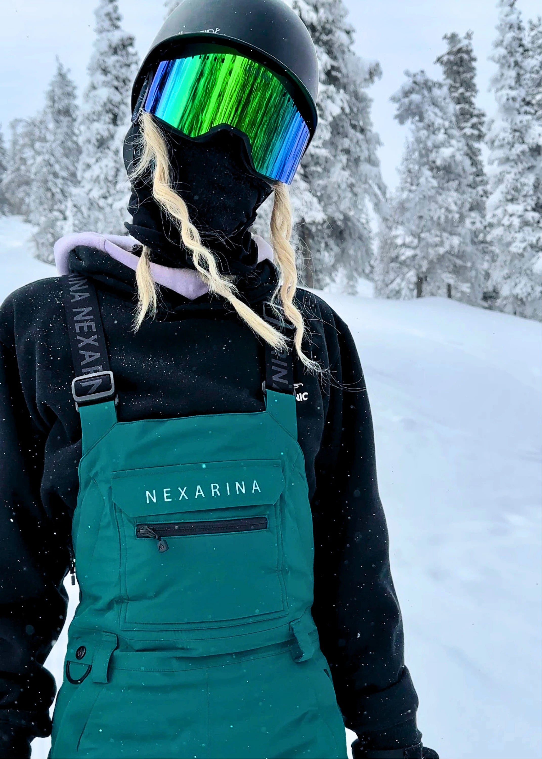 Close-up of snowboarder wearing Nexarina Everette Emerald Green bibs with winter outerwear and goggles.