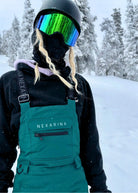 Close-up of snowboarder wearing Nexarina Everette Emerald Green bibs with winter outerwear and goggles.