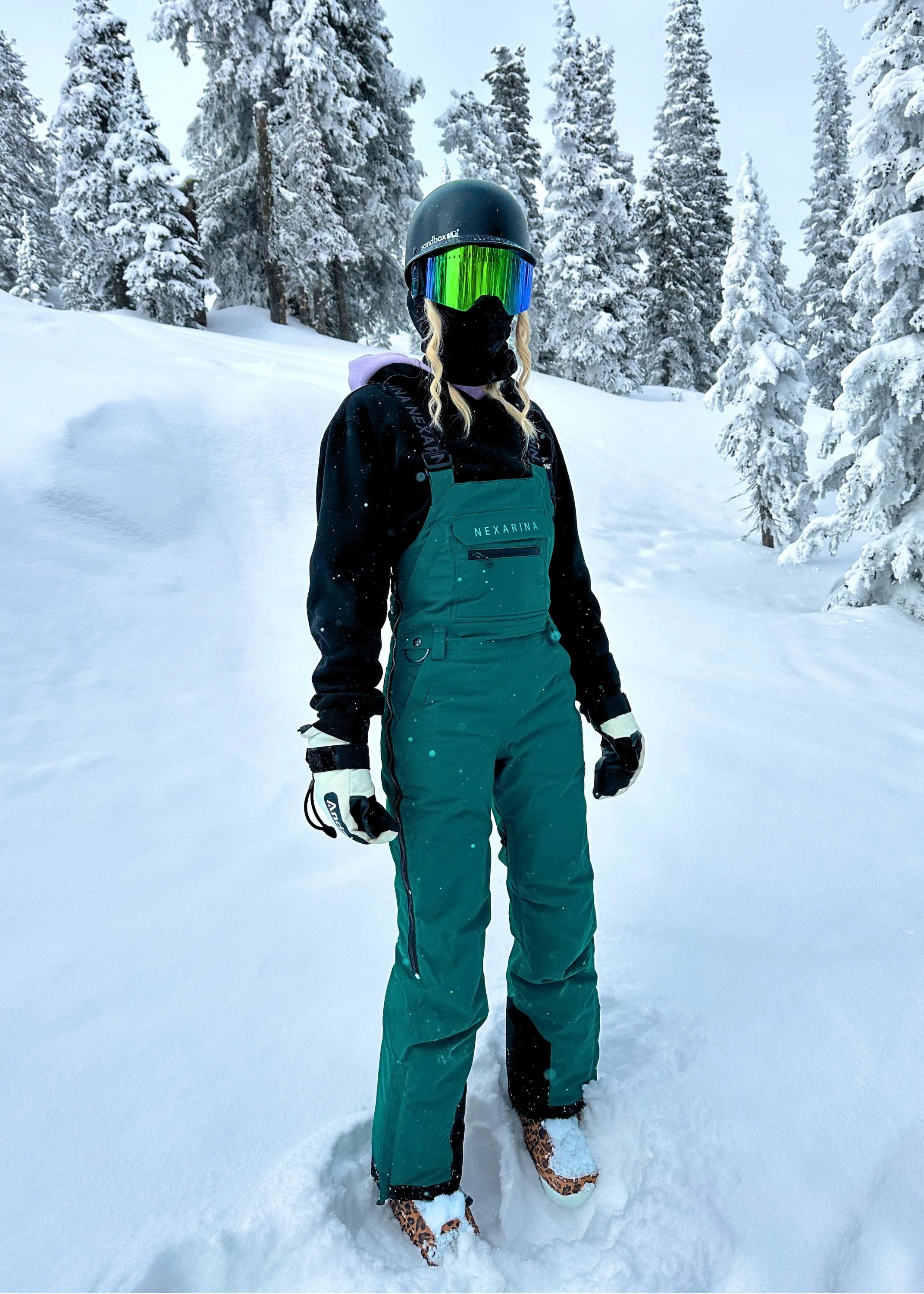 Snowboarder wearing Nexarina Everette Emerald Green insulated bib pants on a snowy mountain trail.