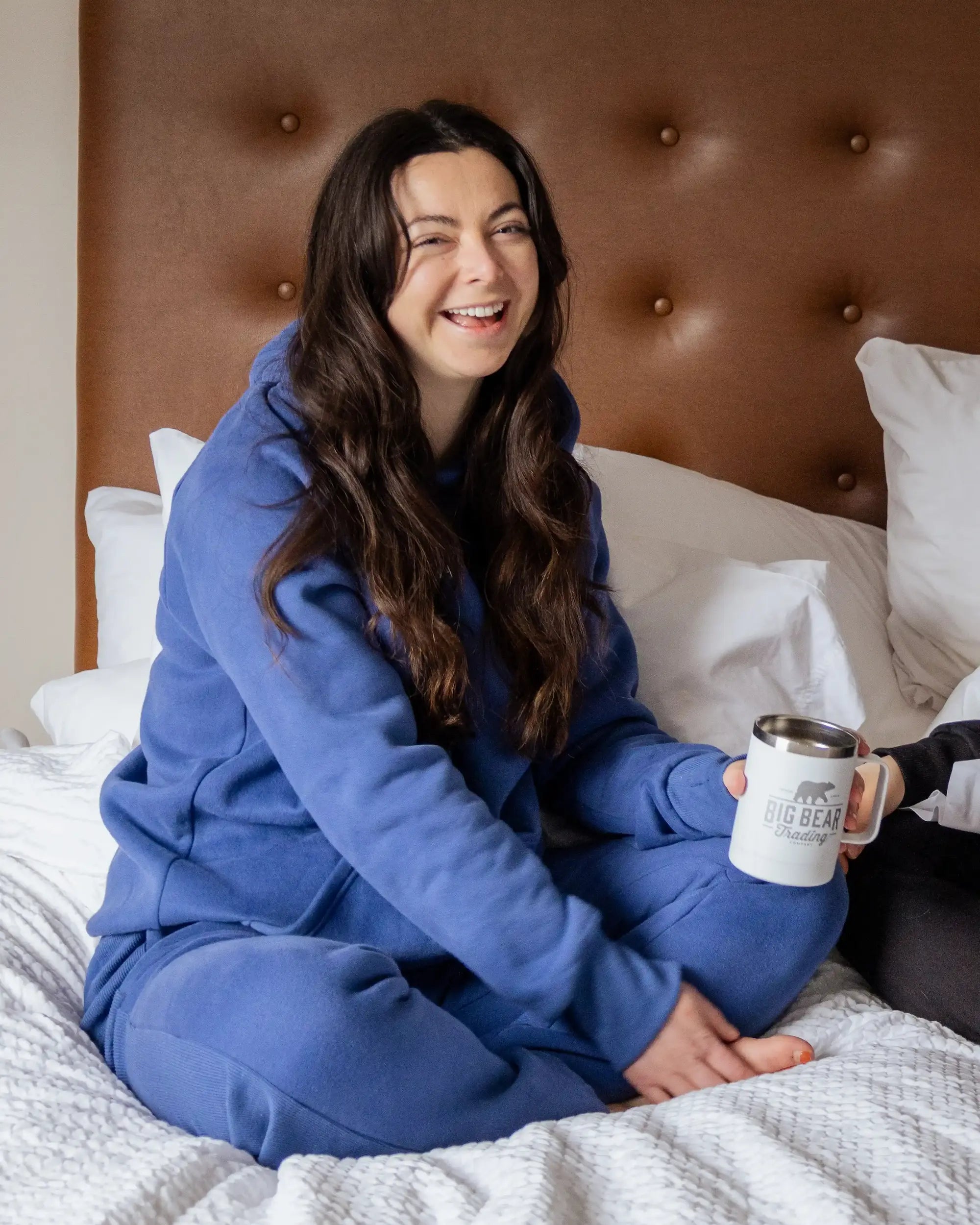 A woman sits cross-legged on a bed in a cozy Everly Blue Nexarina sweatsuit, smiling and holding a Big Bear Trading Co. mug. The mood is relaxed and joyful.
