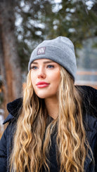 Close-up of a woman wearing a grey Nexarina Emery Beanie, set against a winter forest backdrop.