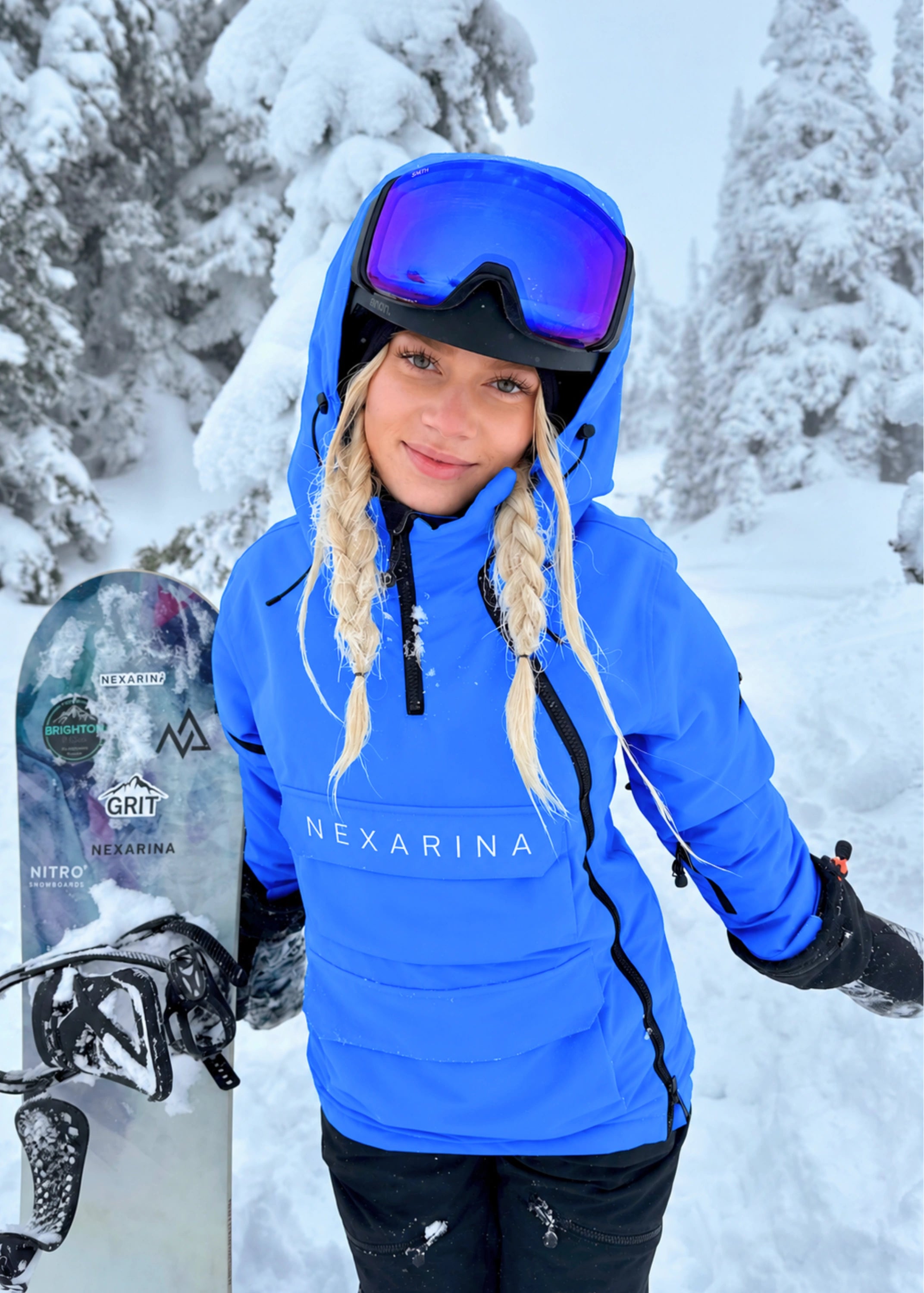 Woman wearing Nexarina Indy Arctic Blue ski jacket with the hood over a snowboard helmet in a snowy forest.