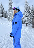 Snowboarder wearing Nexarina Indy Arctic Blue insulated ski jacket with matching snow pants in a snow-covered mountain forest.
