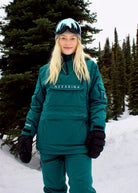 Front view of woman wearing Nexarina Indy Emerald Green women's insulated ski jacket in a snowy mountain setting.