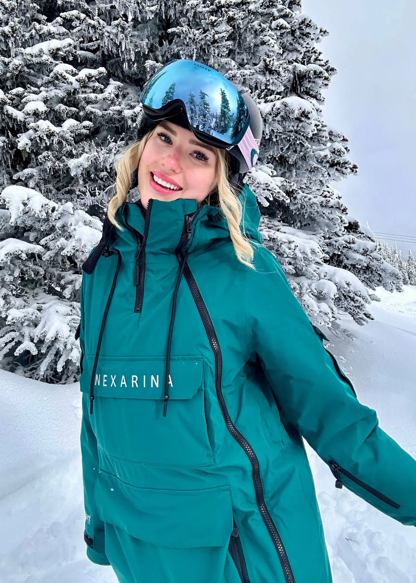 Person wearing a emerald green Nexarina snowsuit with a helmet and goggles in a snowy forest.