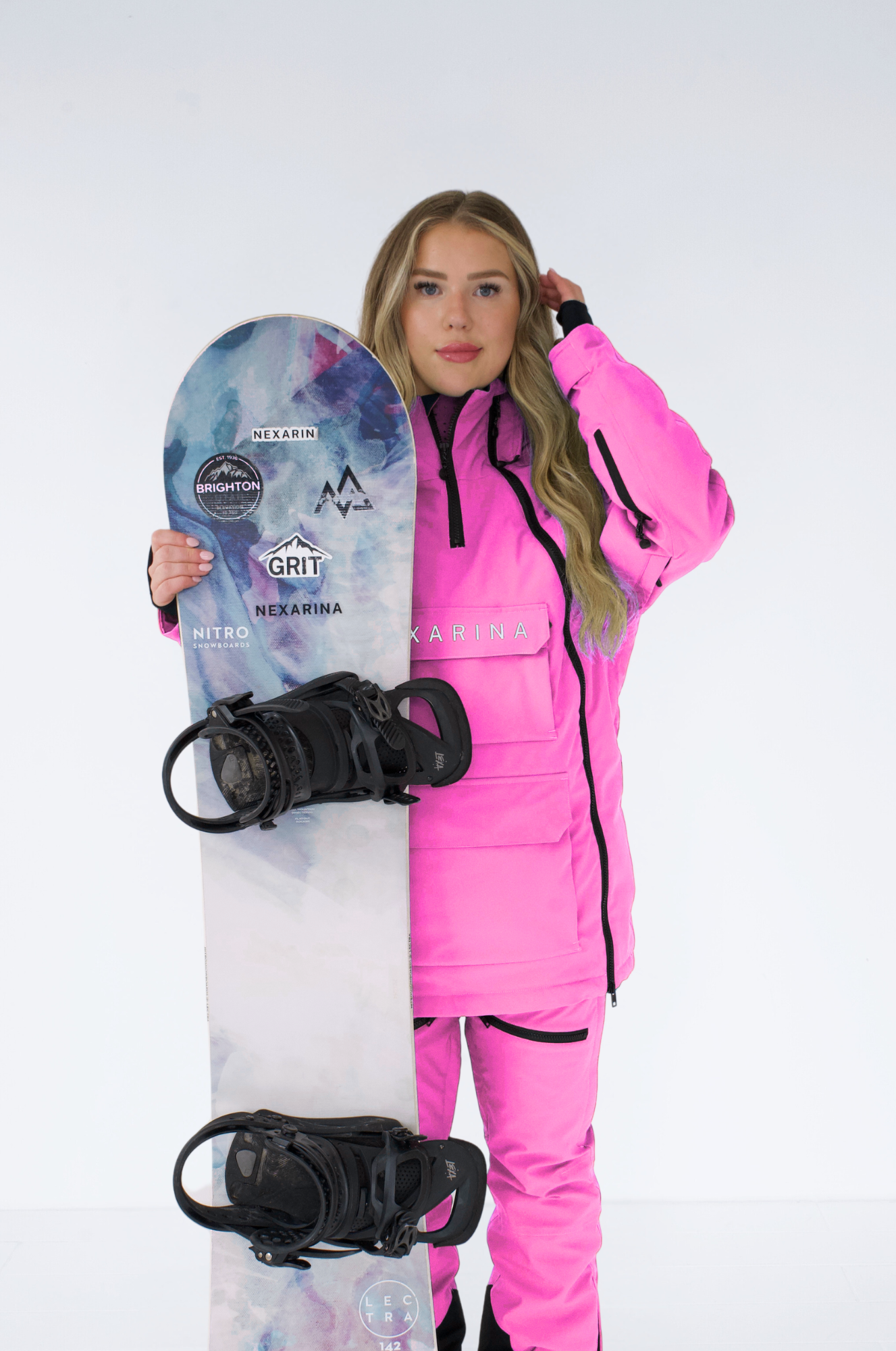 Woman in a pink Nexarina snowboard jacket holding a snowboard, posed against a white backdrop