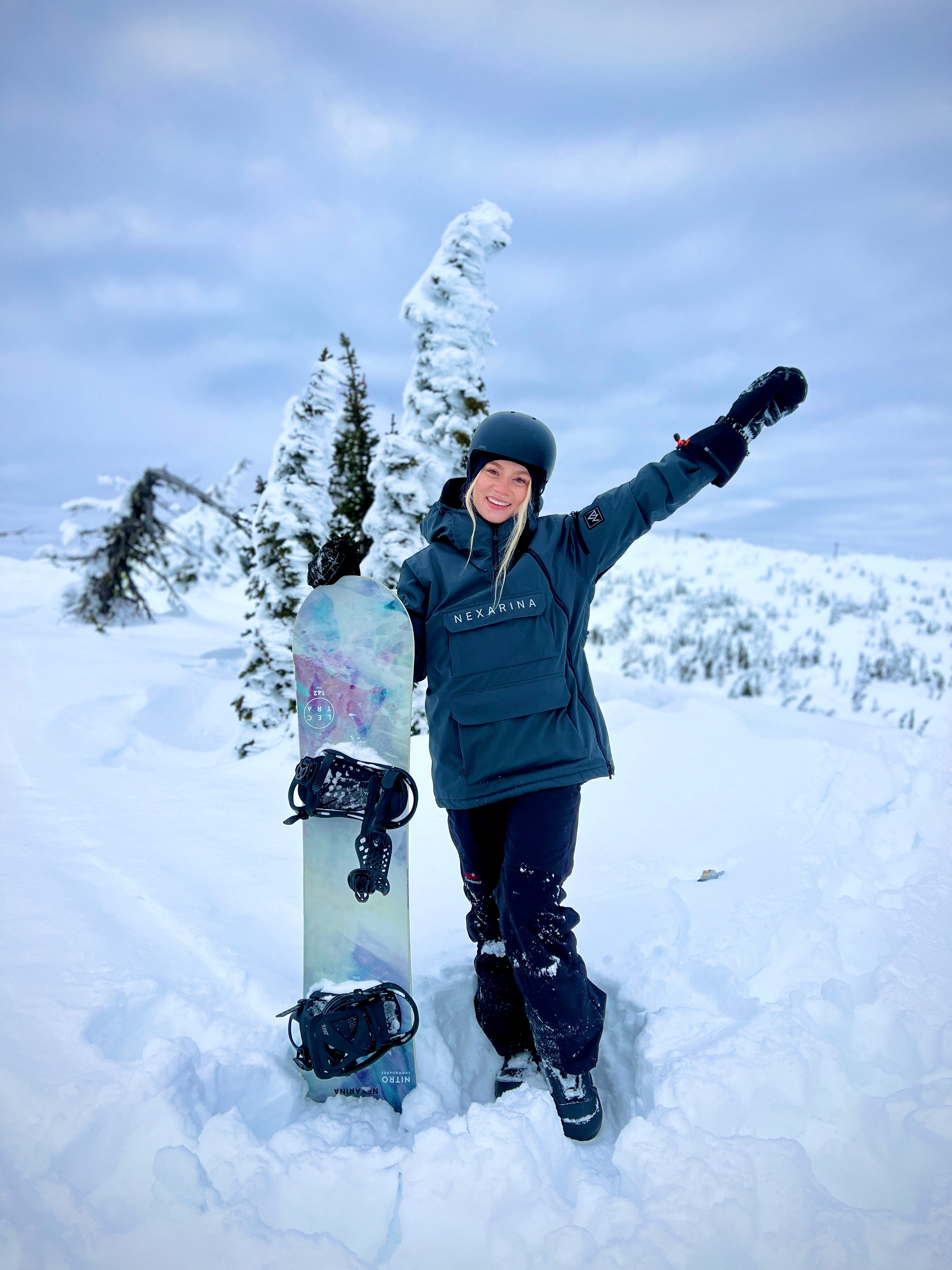 A female snowboarder in a grey Nexarina jacket stands on a snowy mountain, smiling beside her snowboard. Nexarina outerwear is designed for fit and performance