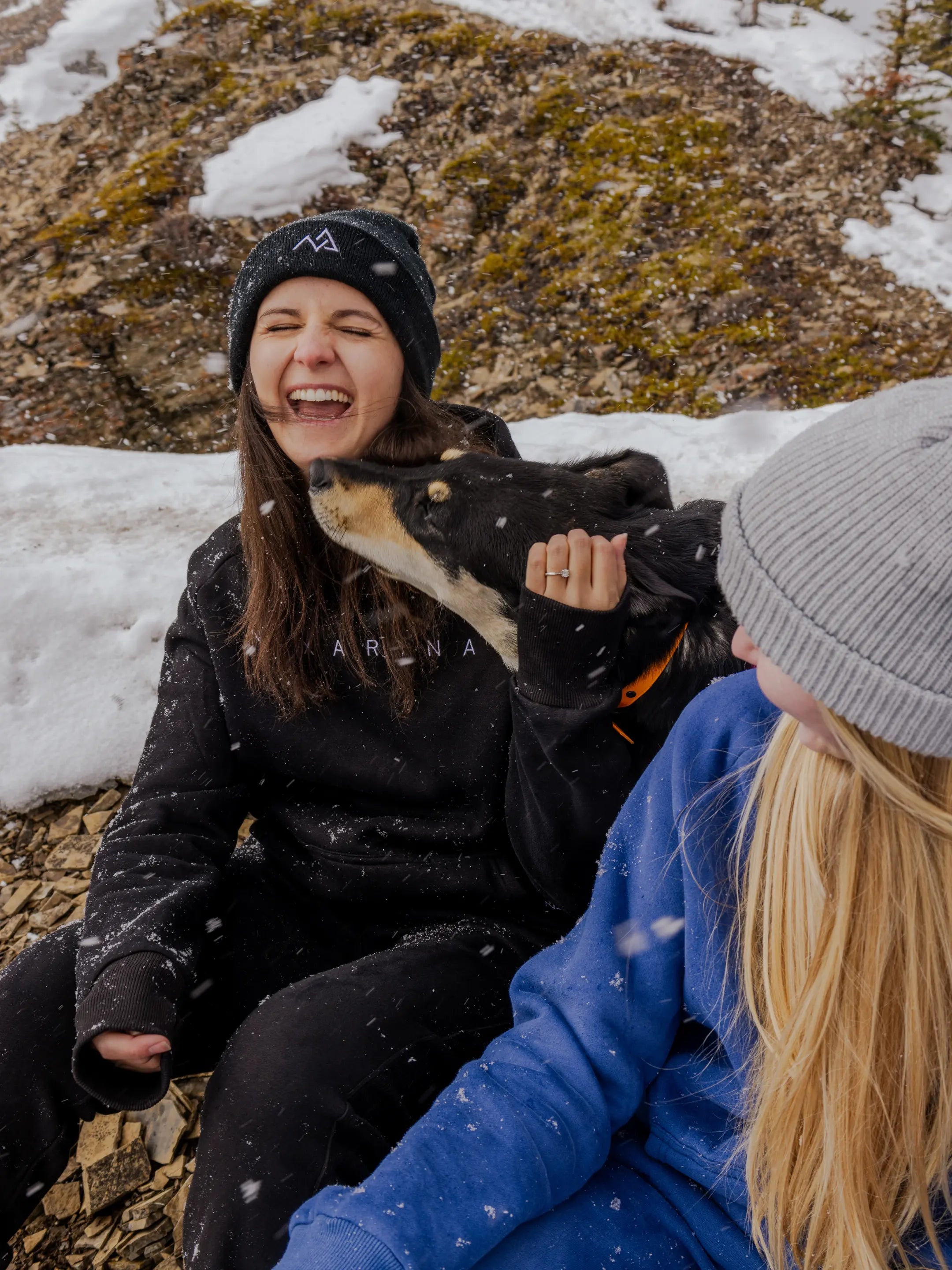 Two women wearing Nexarina beanies and sweatsuits sitting on a snowy mountainside with a dog. One woman laughs joyfully as the dog playfully nuzzles her face.