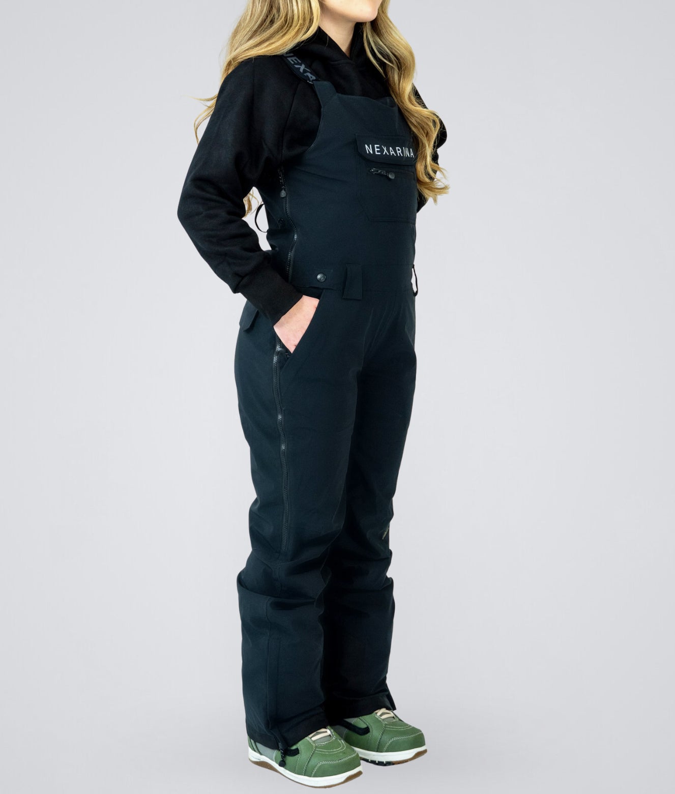 Woman wearing Nexarina Everette women’s black bib snow pants, side profile showing tailored fit and full-length leg coverage.